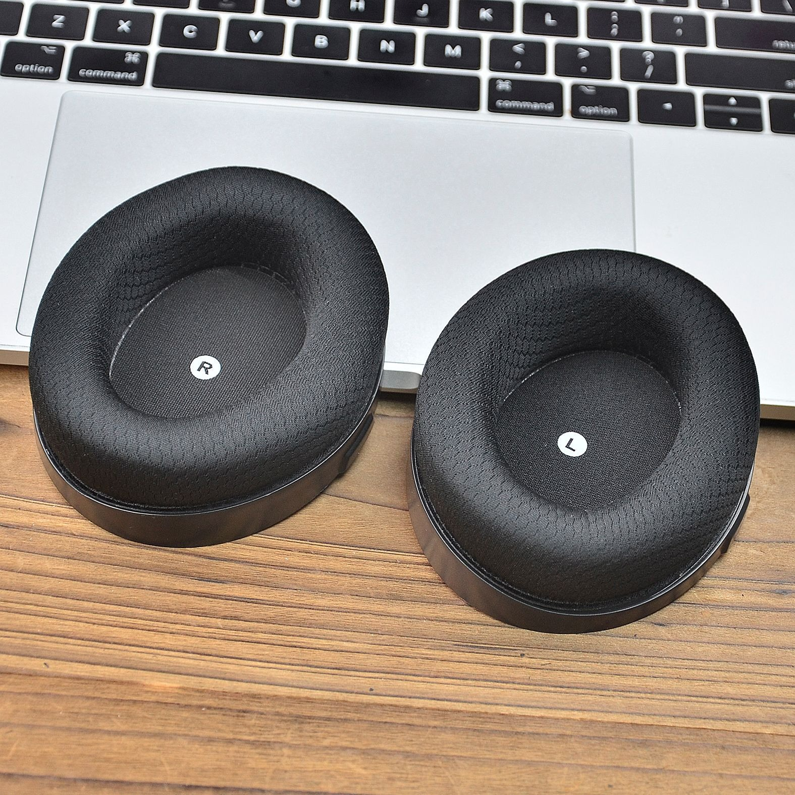Due cuscinetti auricolari neri con etichette 'L' e 'R', posizionati su una tastiera di un laptop e una superficie in legno.