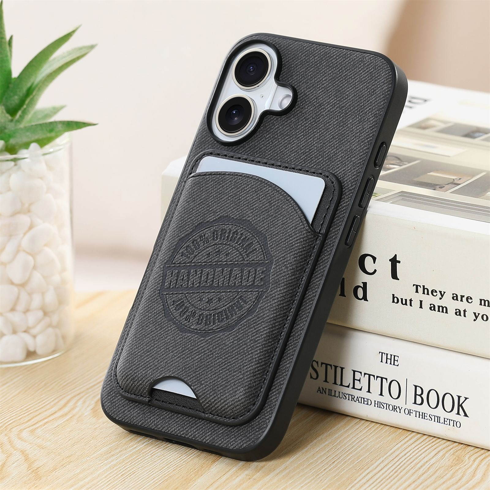 Una custodia grigia per telefono con porta carte. Il supporto ha un logo 'HANDMADE'. Libri sullo sfondo.
