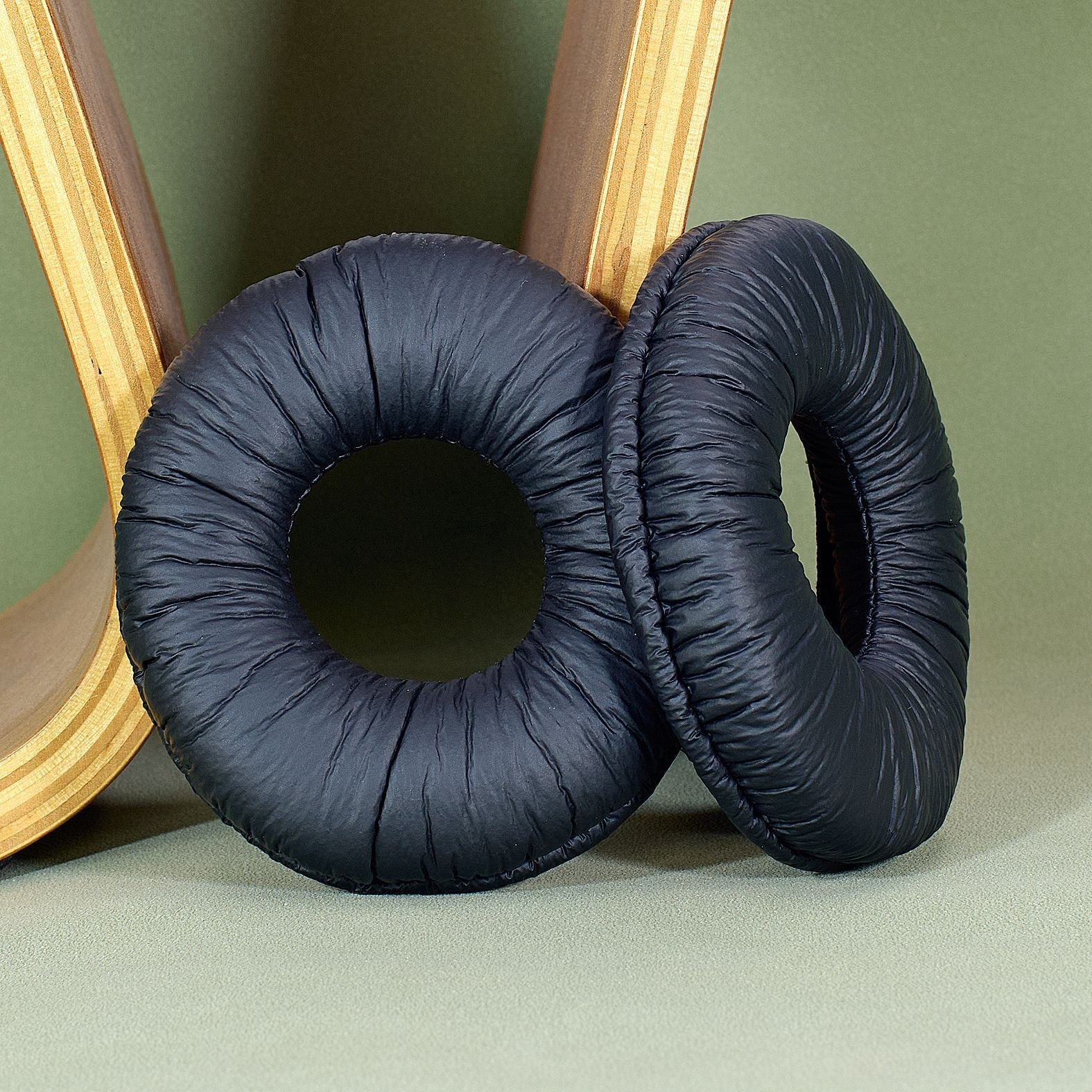 Due cuscinetti auricolari neri e circolari per cuffie sono mostrati su una superficie verde. Sono uno accanto all'altro. Un supporto in legno è sullo sfondo.