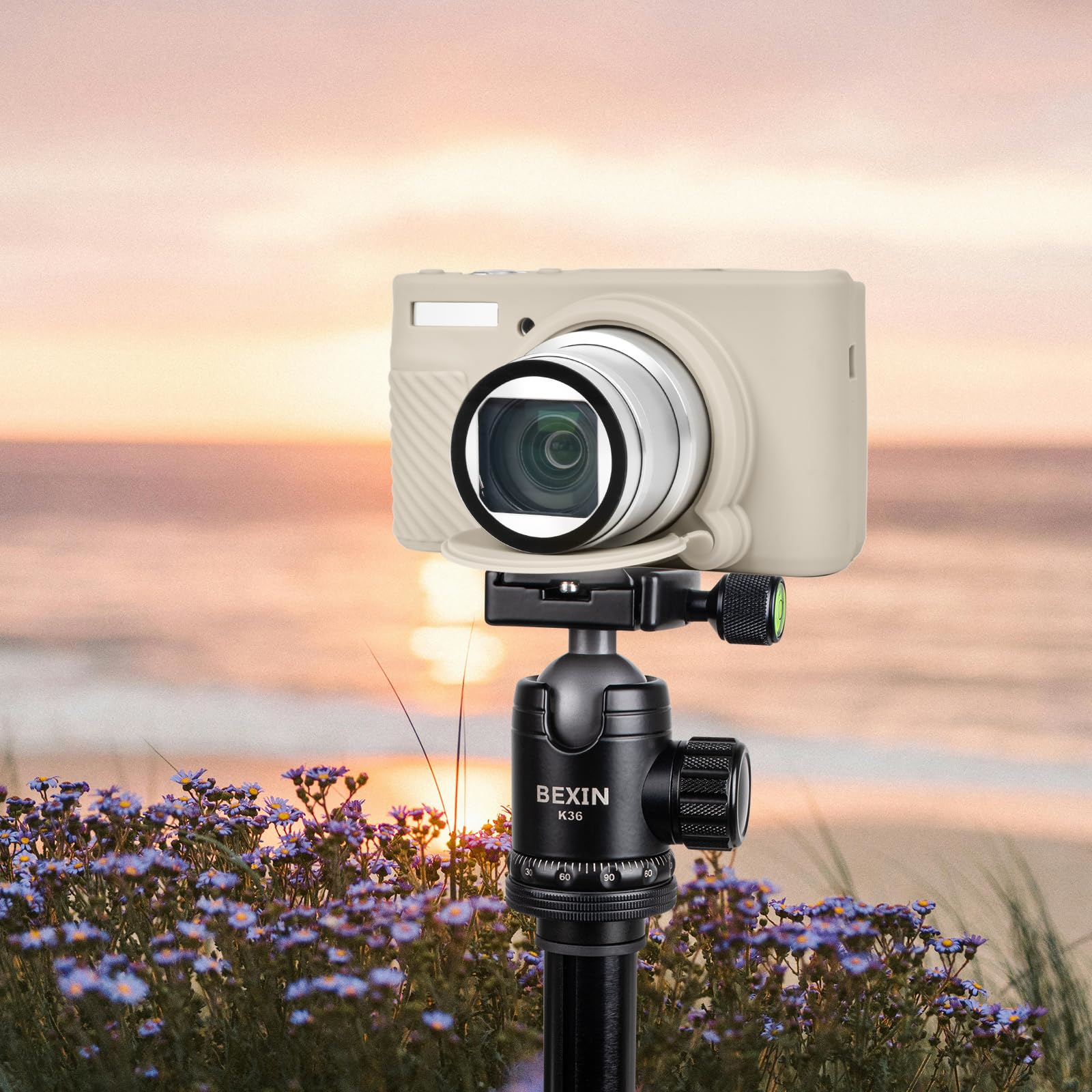 Eine Kamera auf einem Stativ am Strand, mit Sonnenuntergang im Hintergrund und Blumen im Vordergrund.