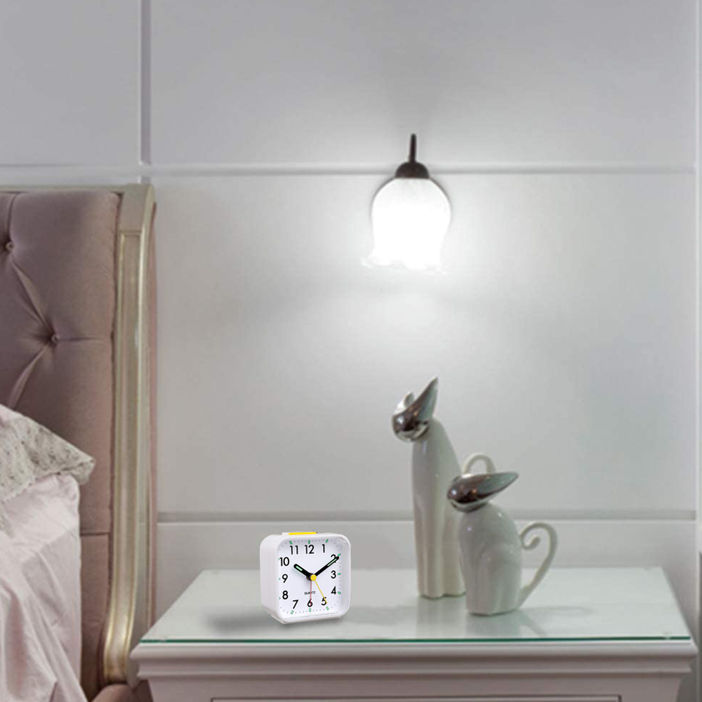Ein weißer Wecker auf einem weißen Tisch neben zwei Katzenfiguren und einer Wandlampe in einem Schlafzimmer.