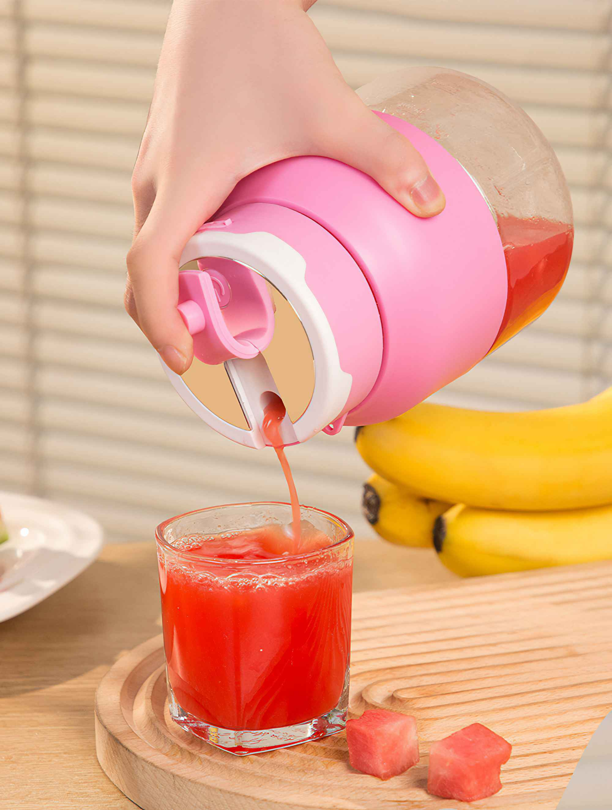 Hand gießt roten Saft aus einem rosa Mixer in ein Glas. Wassermelonenwürfel liegen neben dem Glas.