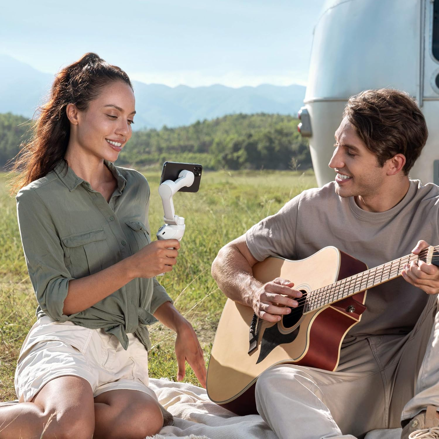Ein Paar sitzt auf einer Decke, die Frau hält ein Telefon auf einem Gimbal, der Mann eine Gitarre.