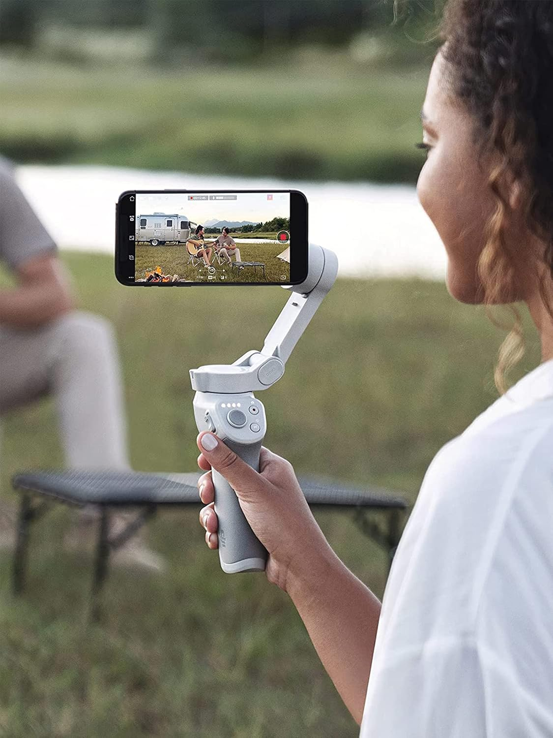 Frau hält Smartphone auf weißem Stabilisator, filmt draußen, mit See und Bäumen im Hintergrund.
