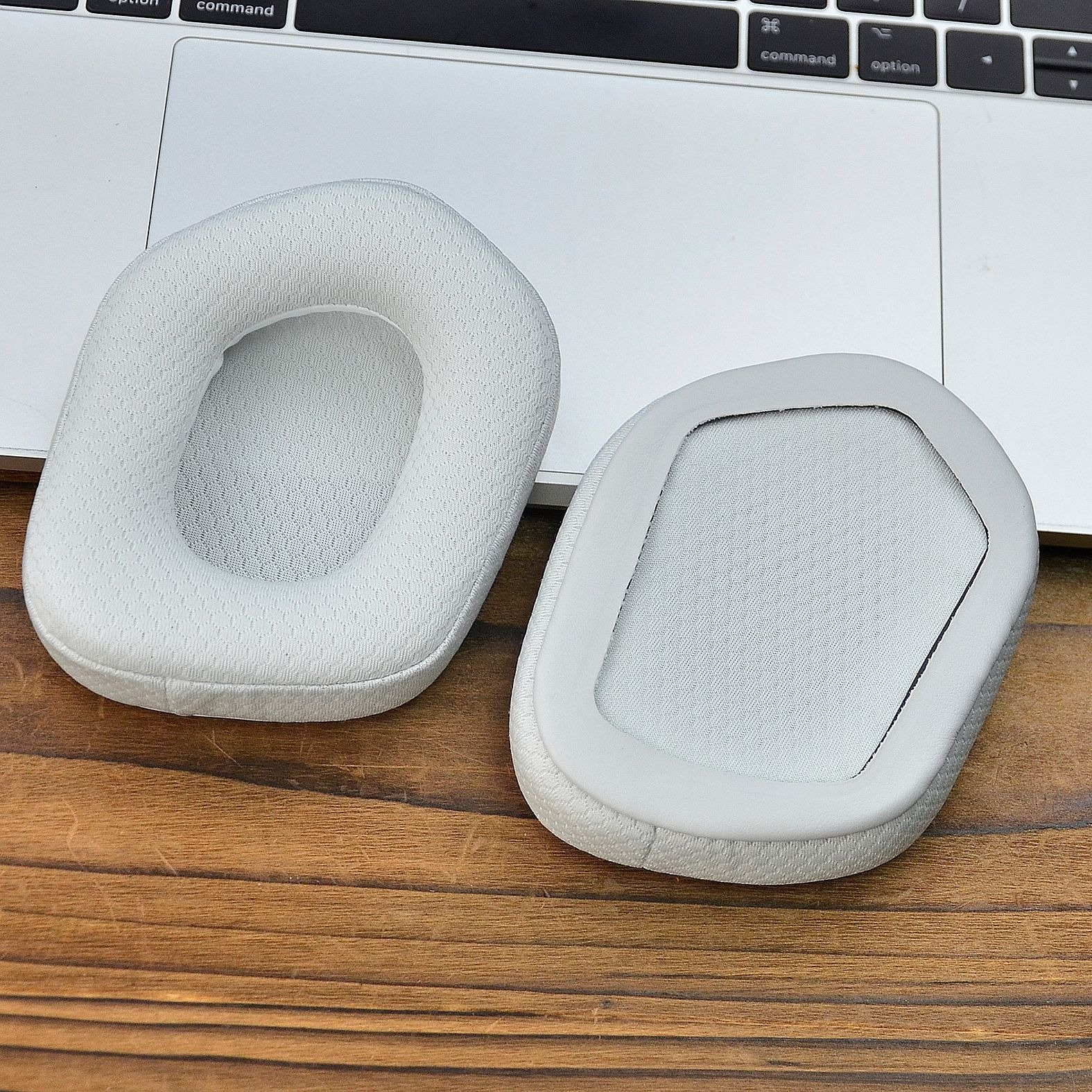Due cuscinetti auricolari grigio chiaro per cuffie, su una superficie di legno, accanto a un laptop.