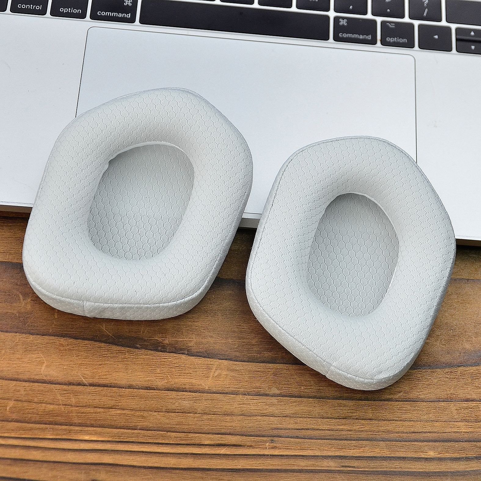 Due cuscinetti auricolari grigio chiaro, a forma ovale, per cuffie, su una superficie di legno con un laptop sullo sfondo.