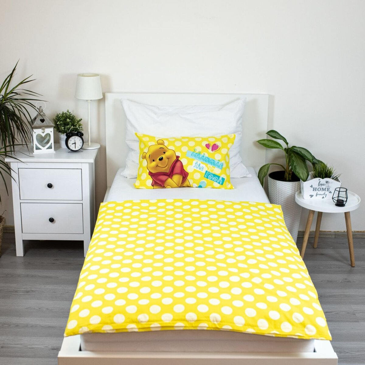 Ein Bett mit gelber Bettwäsche mit Tupfen, einem Winnie-the-Pooh-Kissen und einem weißen Nachttisch.