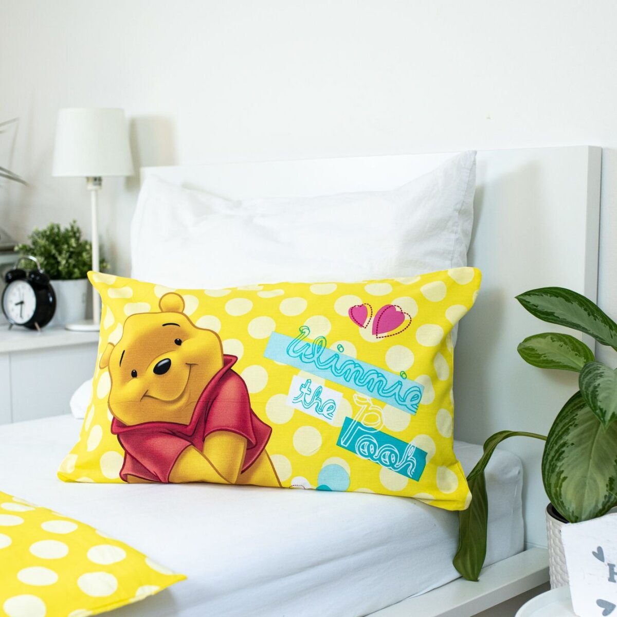 Ein gelbes Winnie-the-Pooh-Kissen auf einem weißen Bett. Es hat ein Tupfenmuster und die Worte Winnie the Pooh.