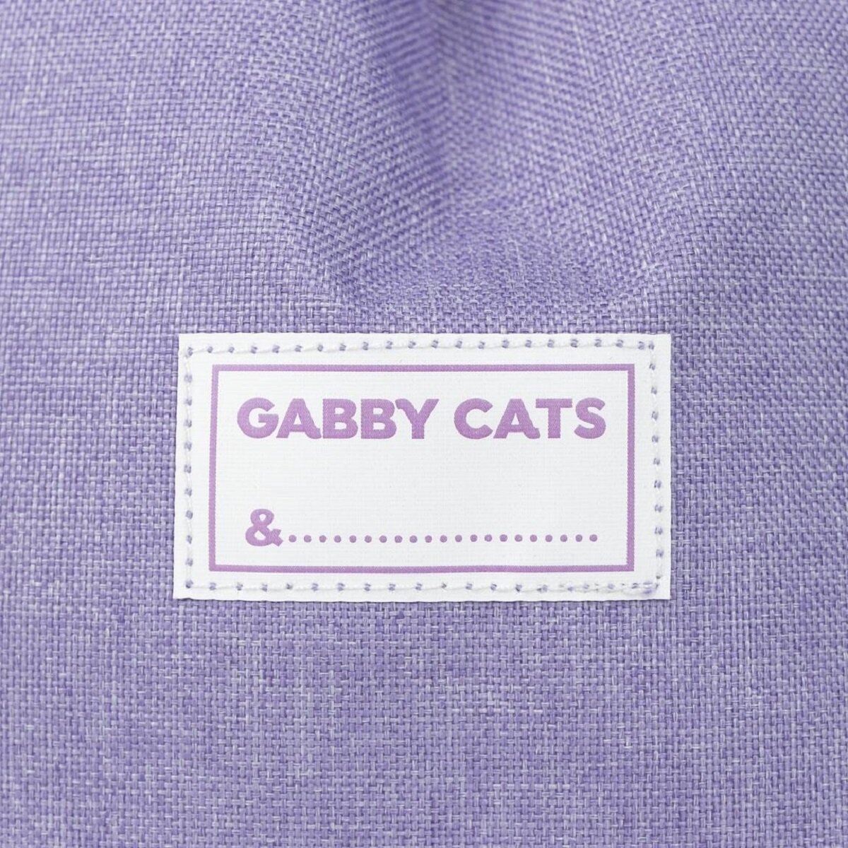 Nahaufnahme eines lila Stoffetiketts mit der Aufschrift 'GABBY CATS &...'. Das Etikett hat eine gepunktete Linie.