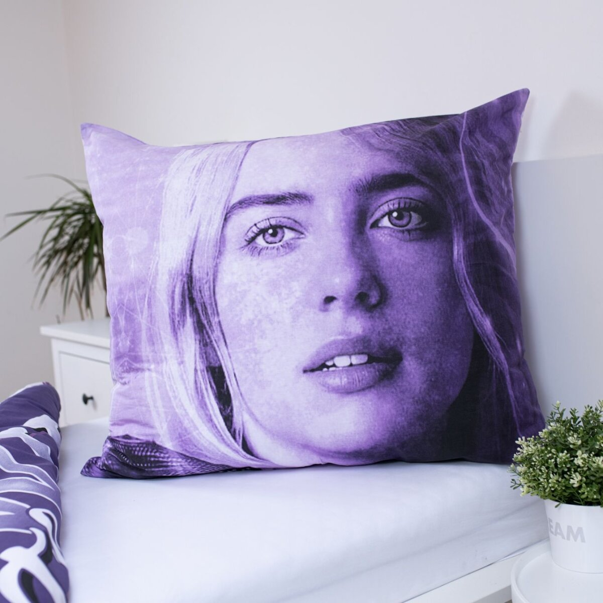 Lila Kissen mit Billie Eilishs Gesicht. Es liegt auf einem Bett. Eine Pflanze und ein weißer Schrank sind im Hintergrund.