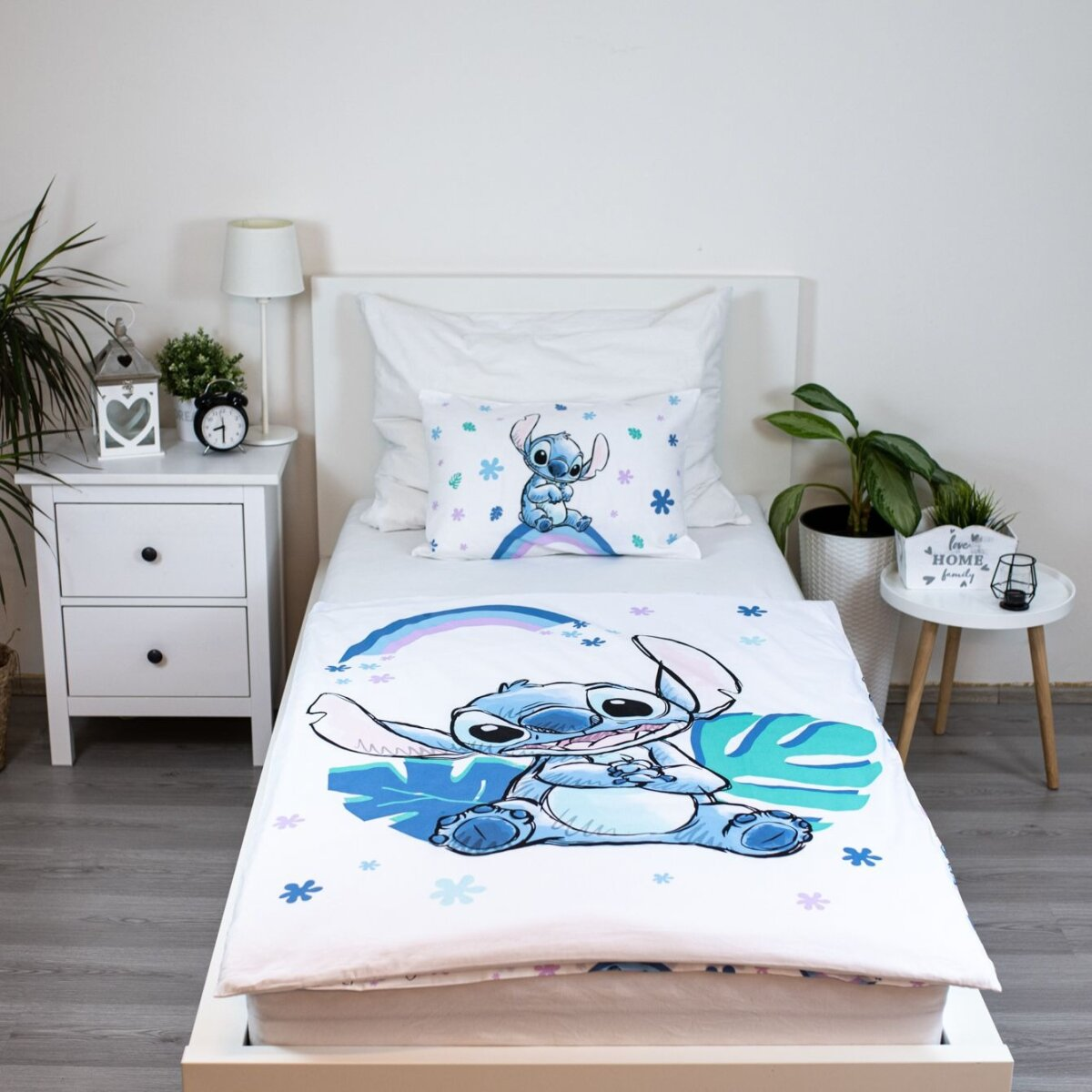 Ein Bett mit einer Stitch-Bettwäsche und einem Kissen. Es gibt einen Nachttisch mit einer Lampe.