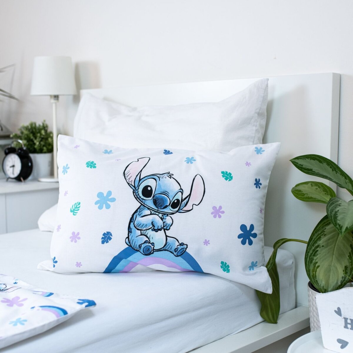 Kissen mit Stitch, blauen Blumen, auf weißem Bett. Nachttisch mit Lampe und Pflanze sichtbar.
