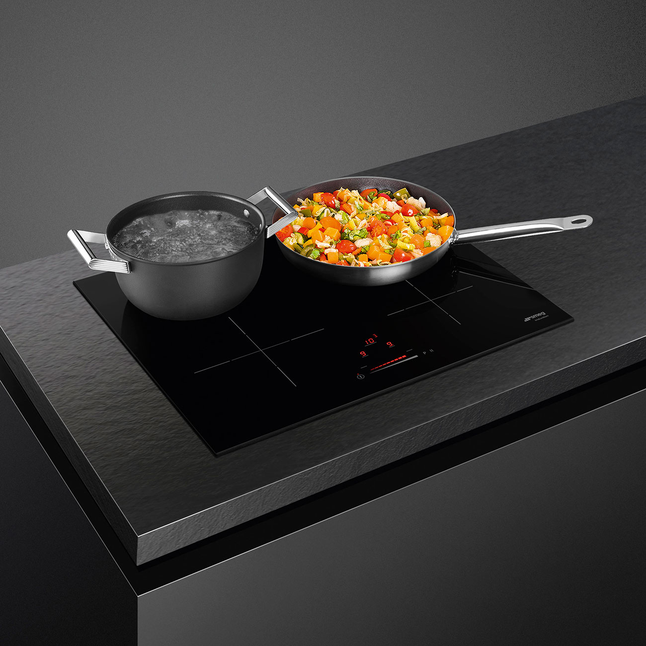 Piano cottura a induzione nero con pentola e padella con cibo, su un piano di lavoro nero con sfondo scuro.