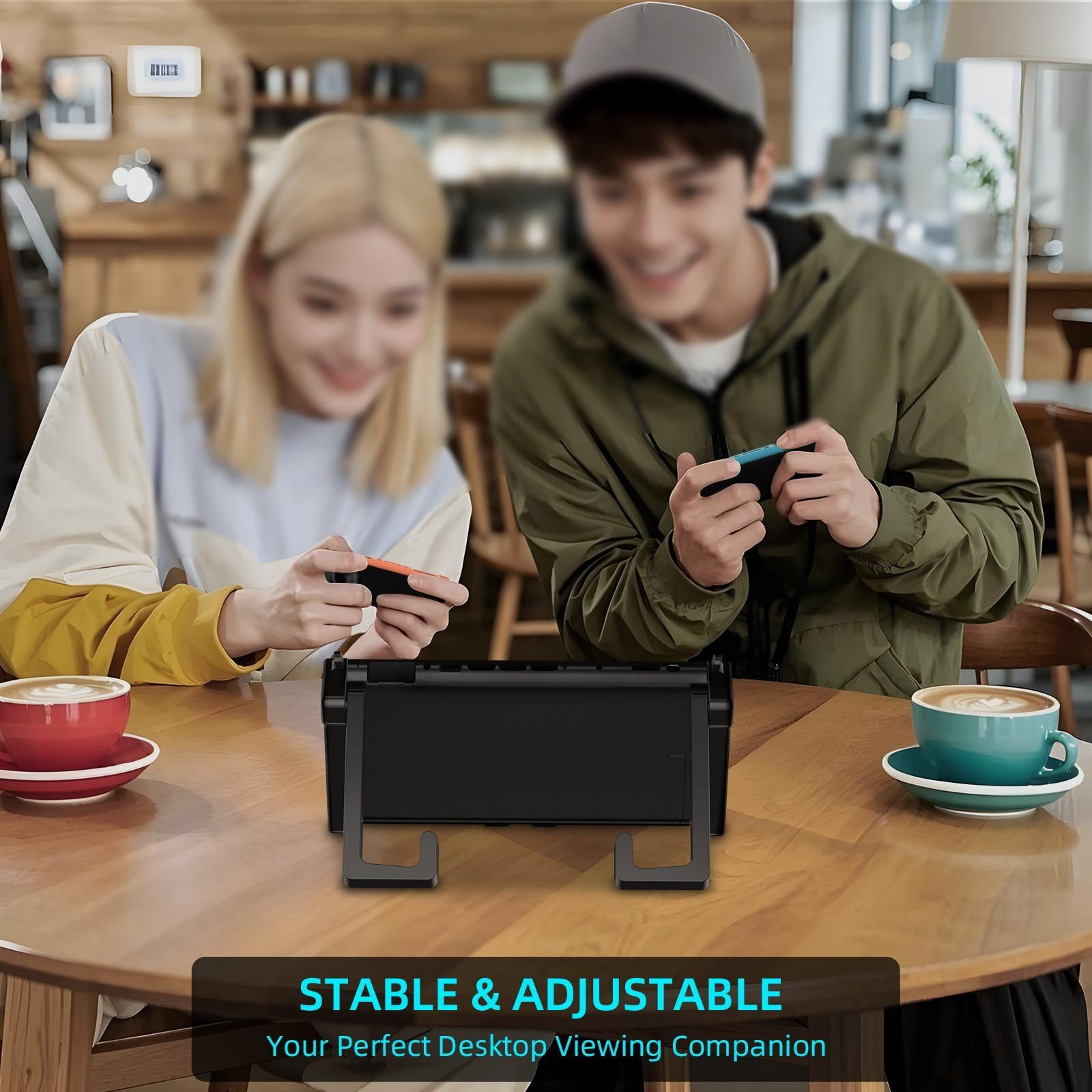 Zwei Personen spielen Videospiele an einem Tisch. Das Spiel ist auf einem Ständer. Kaffeetassen sichtbar.