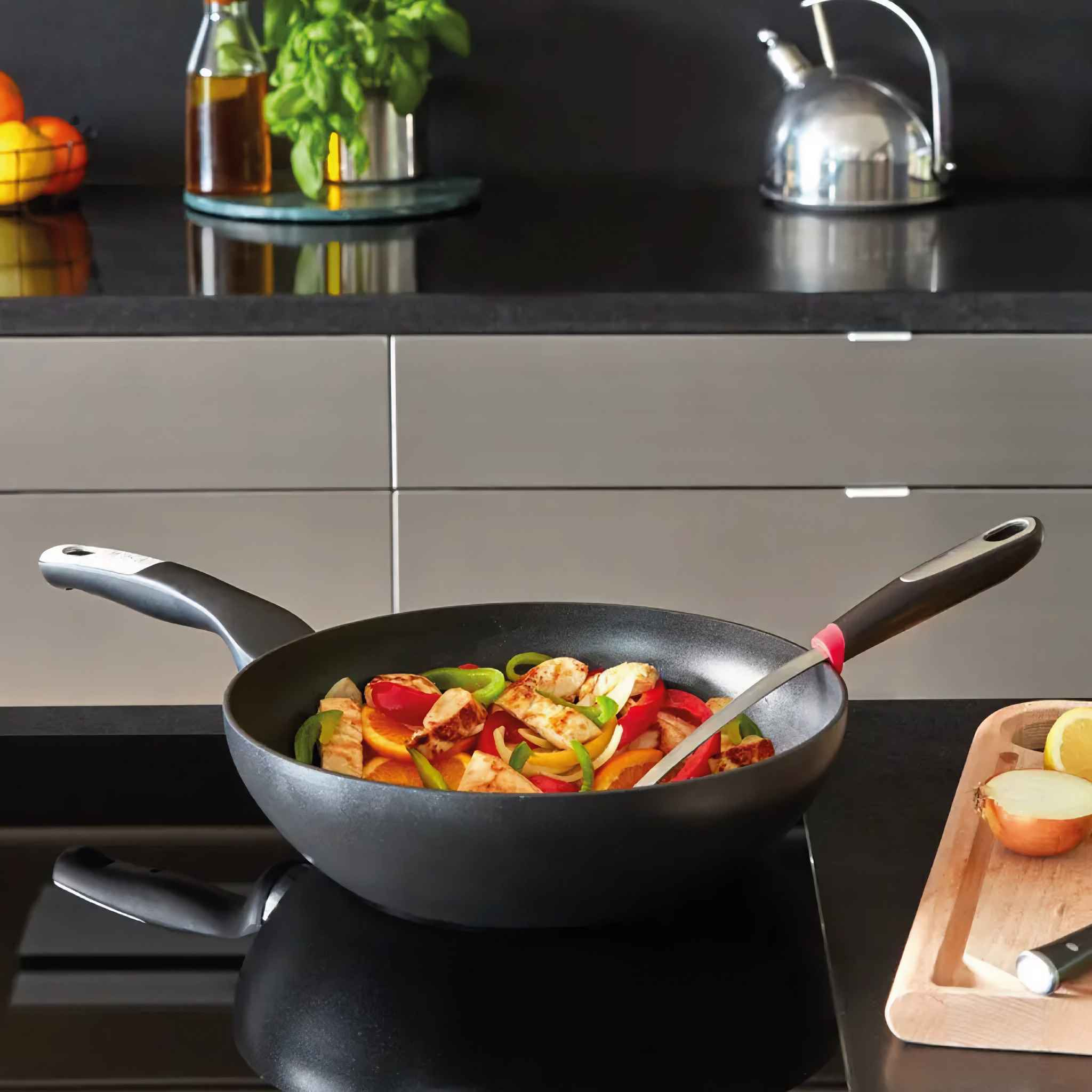 Eine schwarze Wok-Pfanne mit gekochtem Essen, darunter Huhn und Gemüse, steht auf einem schwarzen Herd. Andere Gegenstände sind in der Nähe.