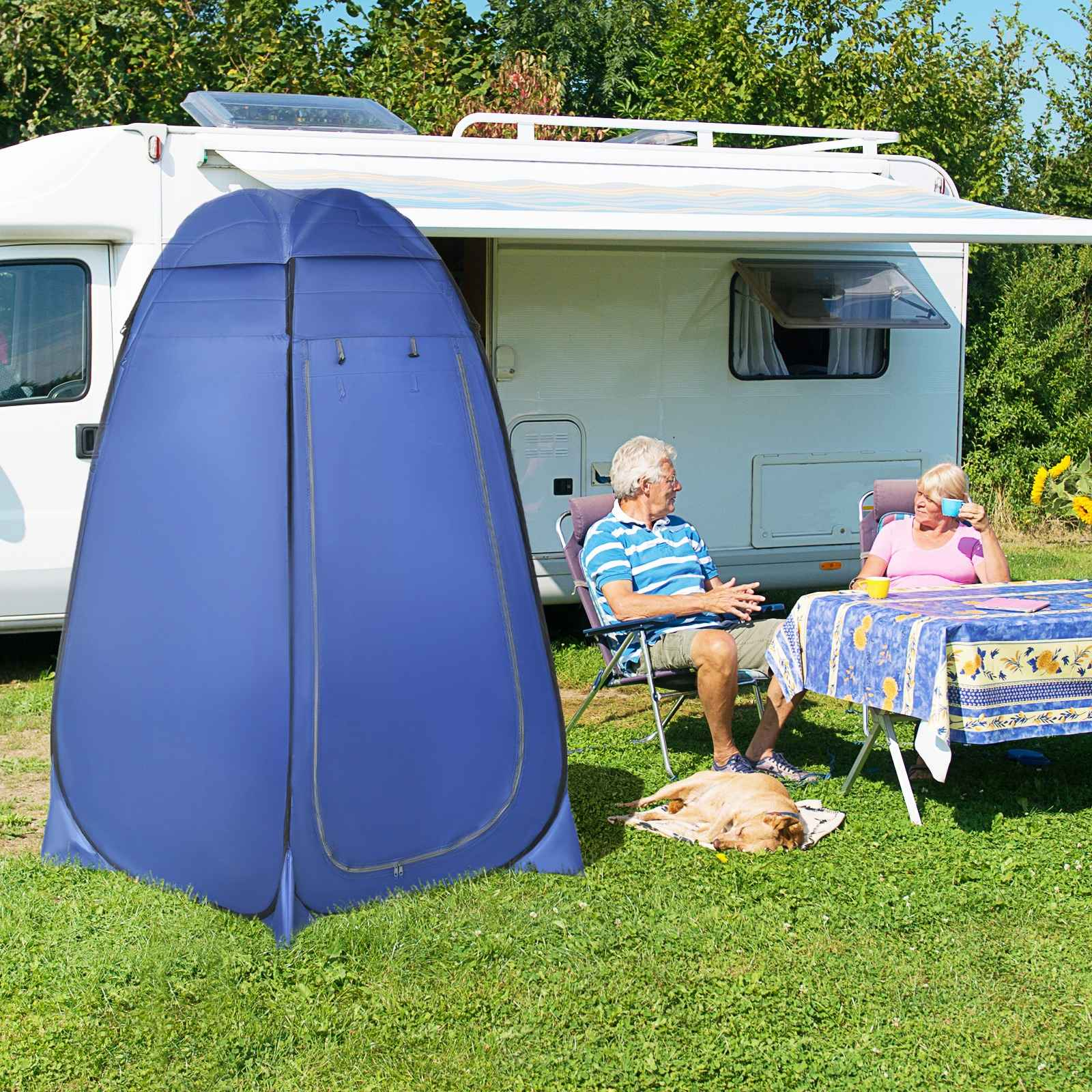 Ein blaues Pop-up-Zelt steht auf einer Wiese. Ein Paar sitzt in der Nähe an einem Tisch. Ein Hund liegt auf dem Gras.