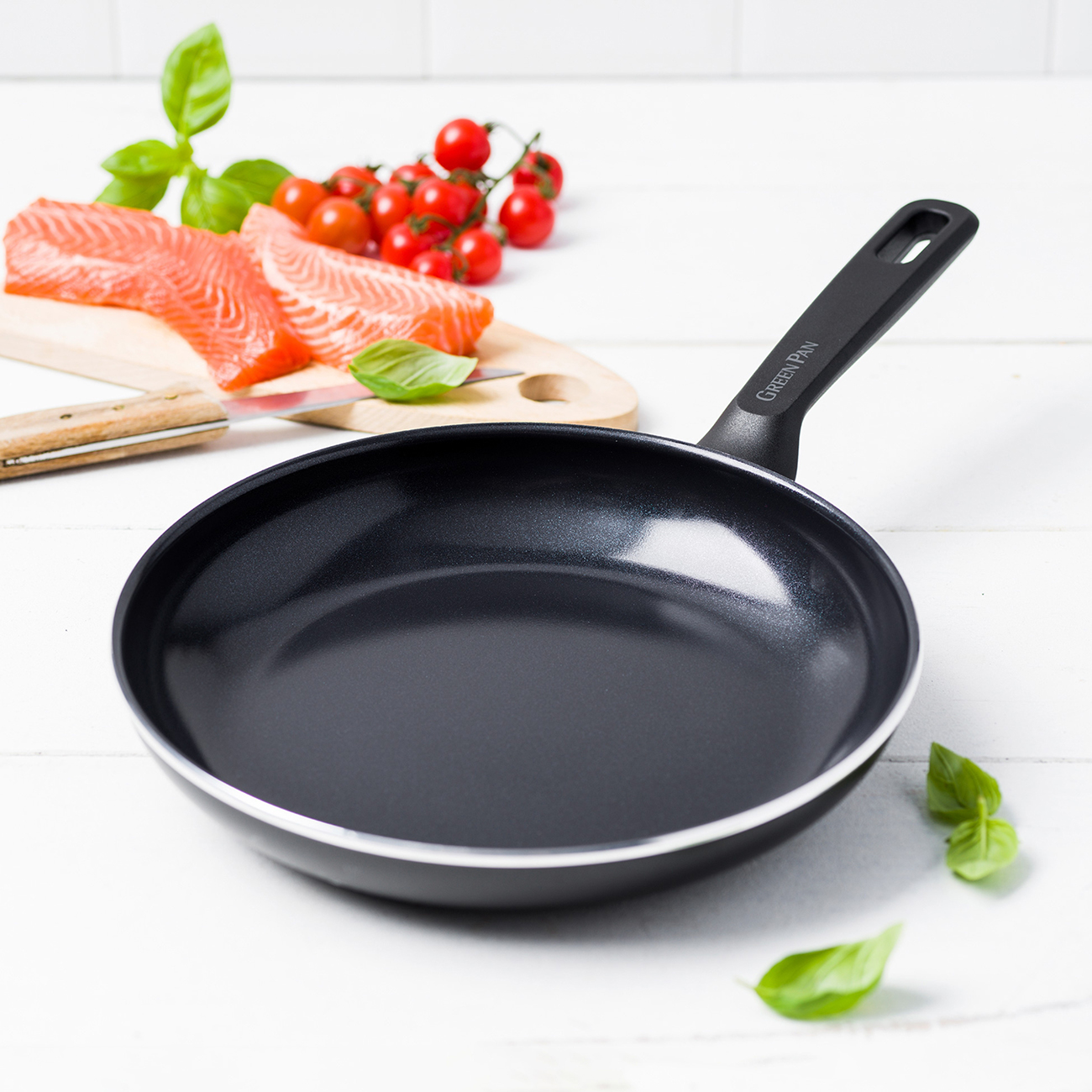 Eine schwarze Bratpfanne auf einem weißen Tisch mit Lachs, Tomaten und Basilikum im Hintergrund.