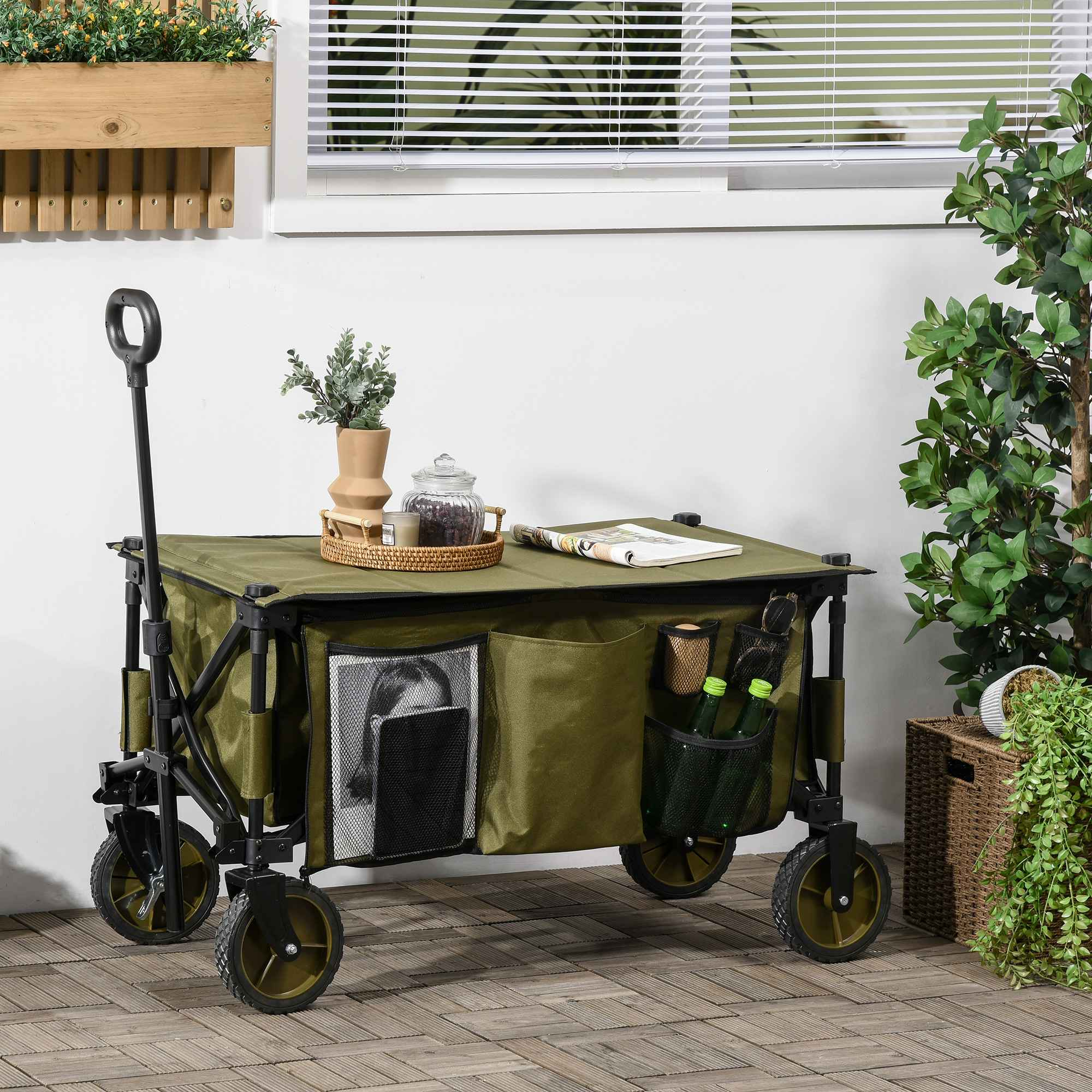 Ein grüner Transportwagen mit schwarzem Rahmen. Der Wagen ist mit Gegenständen gefüllt, in einer Gartensituation, neben Fenster und Pflanzen.