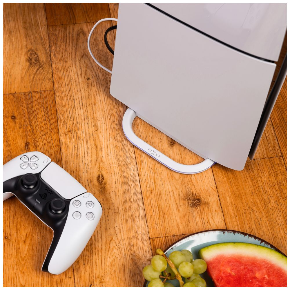 Una console di gioco bianca, controller, uva, fetta di anguria, su un pavimento di legno.