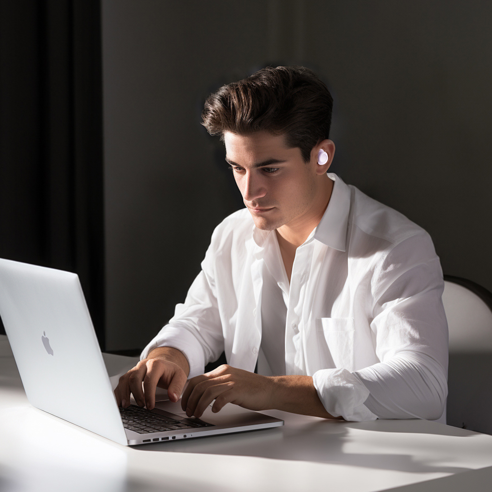 Ein Mann sitzt an einem Tisch und benutzt einen Laptop. Er hat einen Ohrhörer im Ohr. Dunkler Hintergrund.