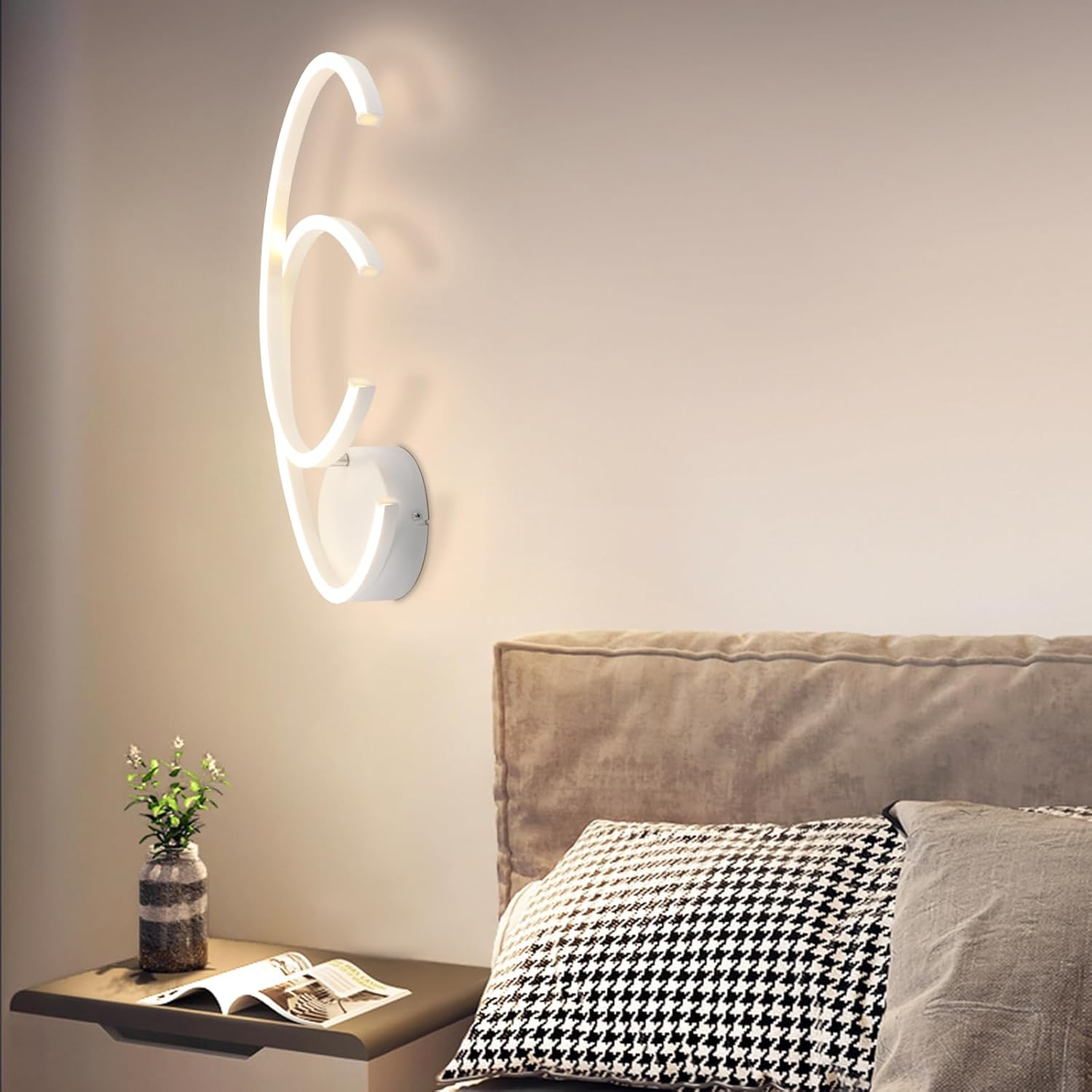 Lampada da parete bianca in una camera da letto. La lampada è accesa, con un letto e un comodino con un libro in primo piano.