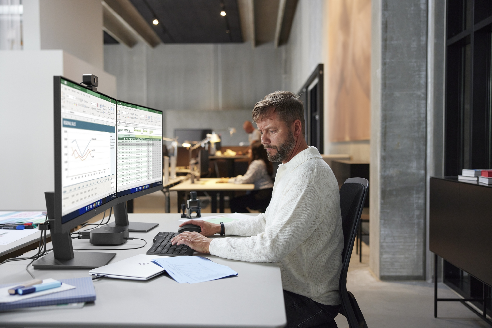 Mężczyzna pracuje przy biurku z dwoma monitorami. W tle ludzie i betonowa ściana.