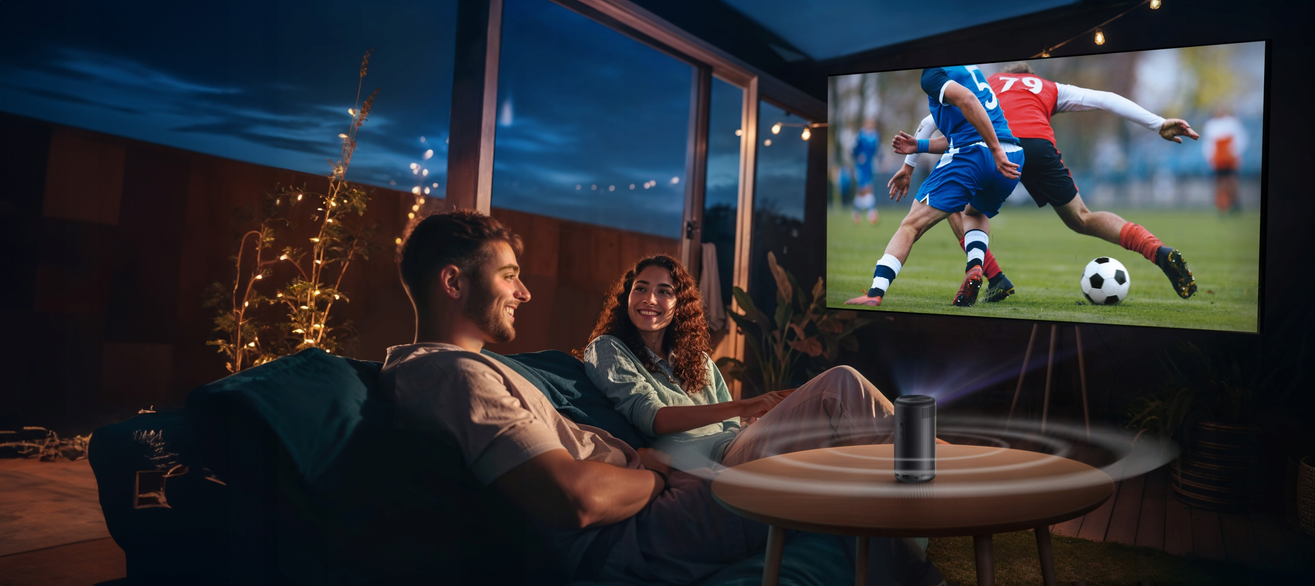Para ogląda mecz piłki nożnej na ekranie. Głośnik stoi na stole.