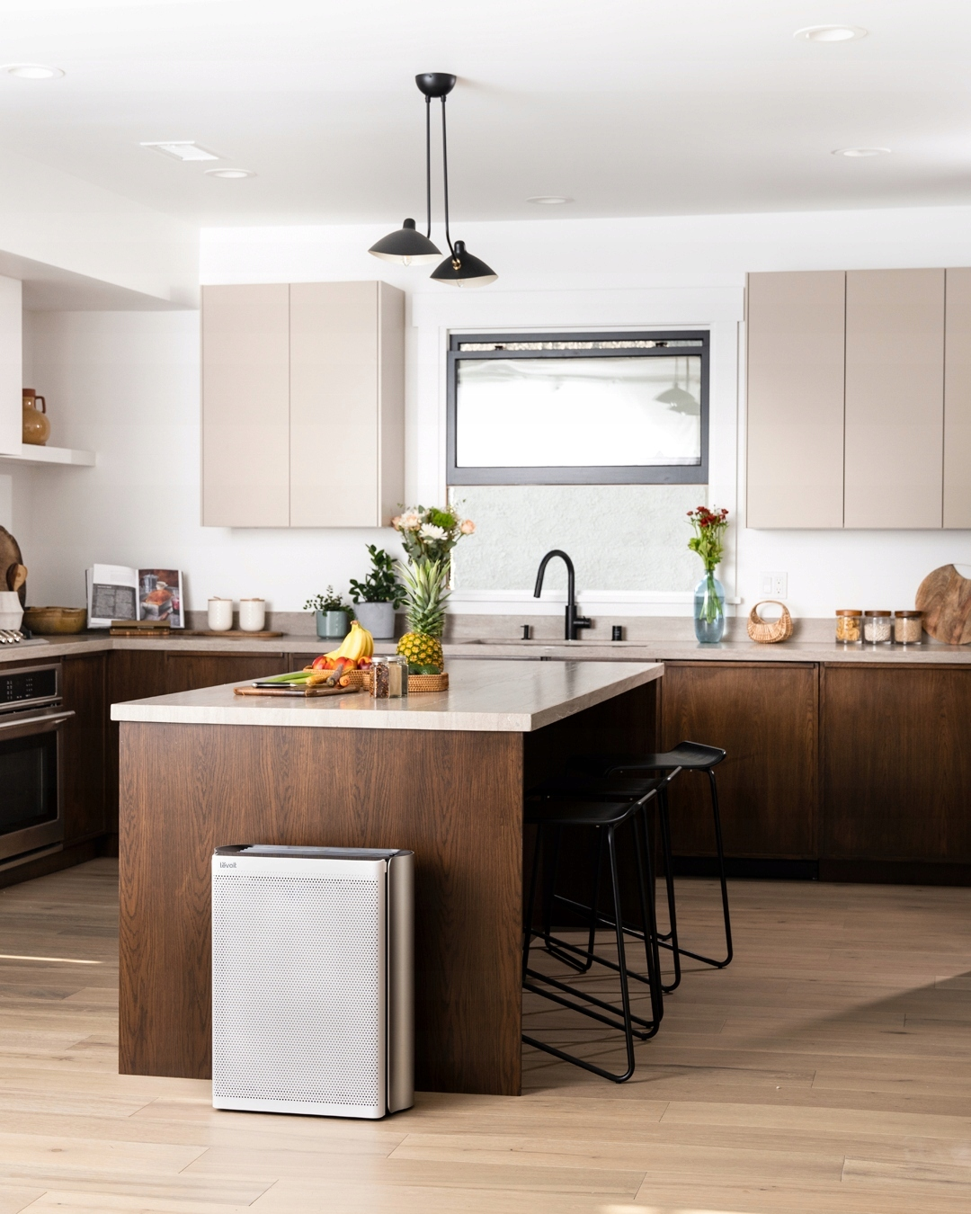 Kuchnia z białym oczyszczaczem powietrza. Wyspa kuchenna z owocami, kwiatami i stołkami. Drewniane szafki i podłogi.