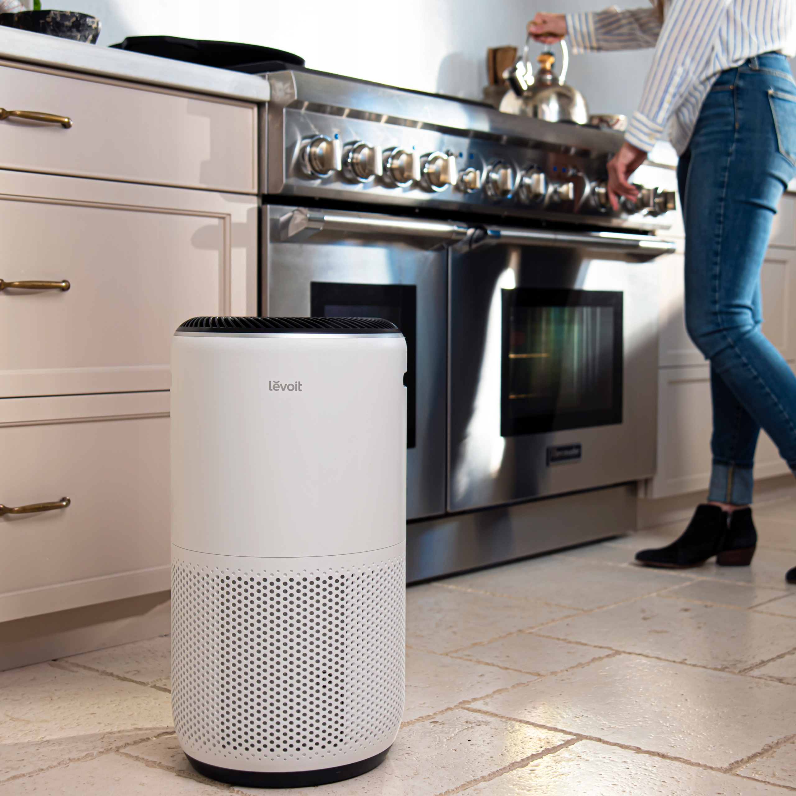Biały oczyszczacz powietrza Levoit w kuchni. Kobieta stoi w tle w pobliżu kuchenki.