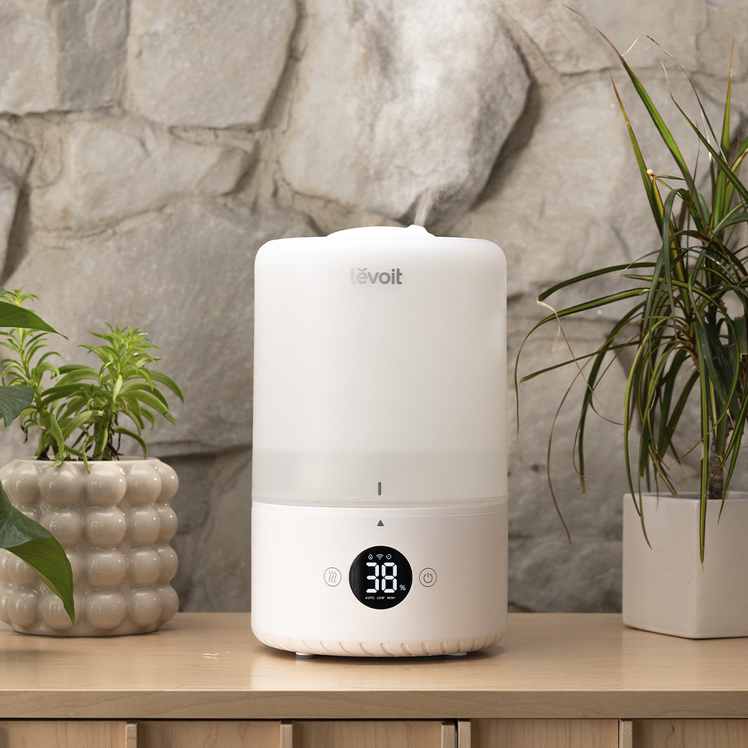 Biały nawilżacz powietrza stoi na drewnianym stole z roślinami. Wyświetla cyfrową temperaturę.