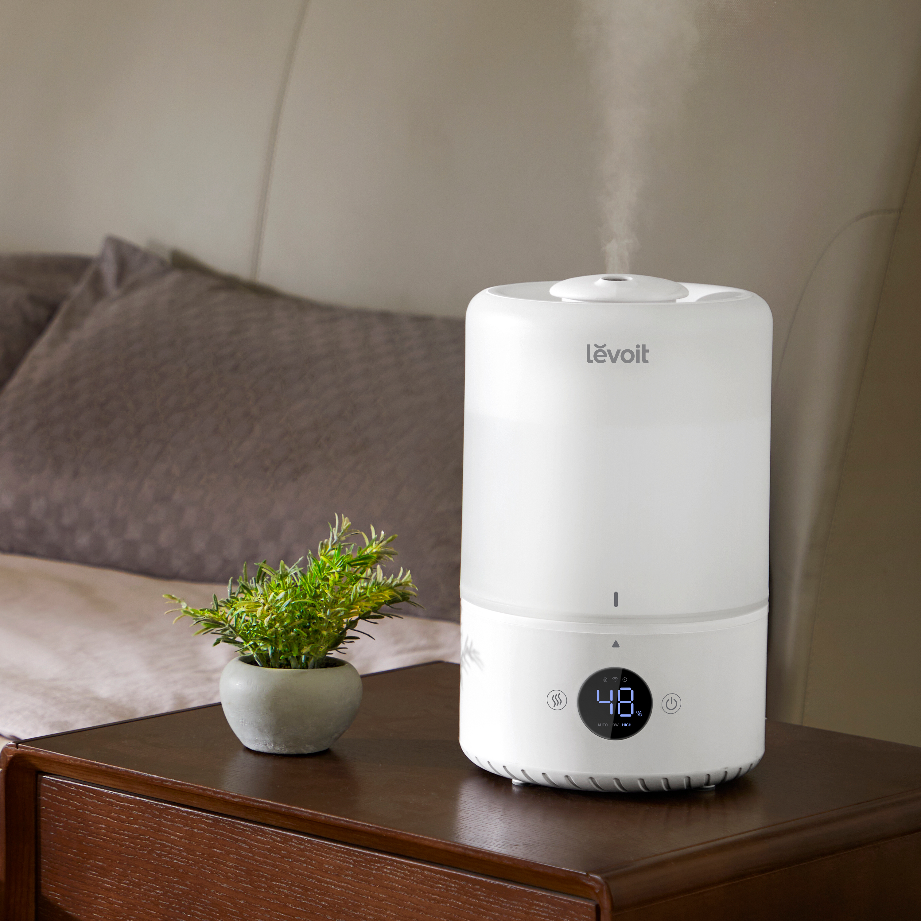 Biały nawilżacz powietrza stoi na stoliku nocnym z małą rośliną. Para jest widoczna. W tle łóżko.
