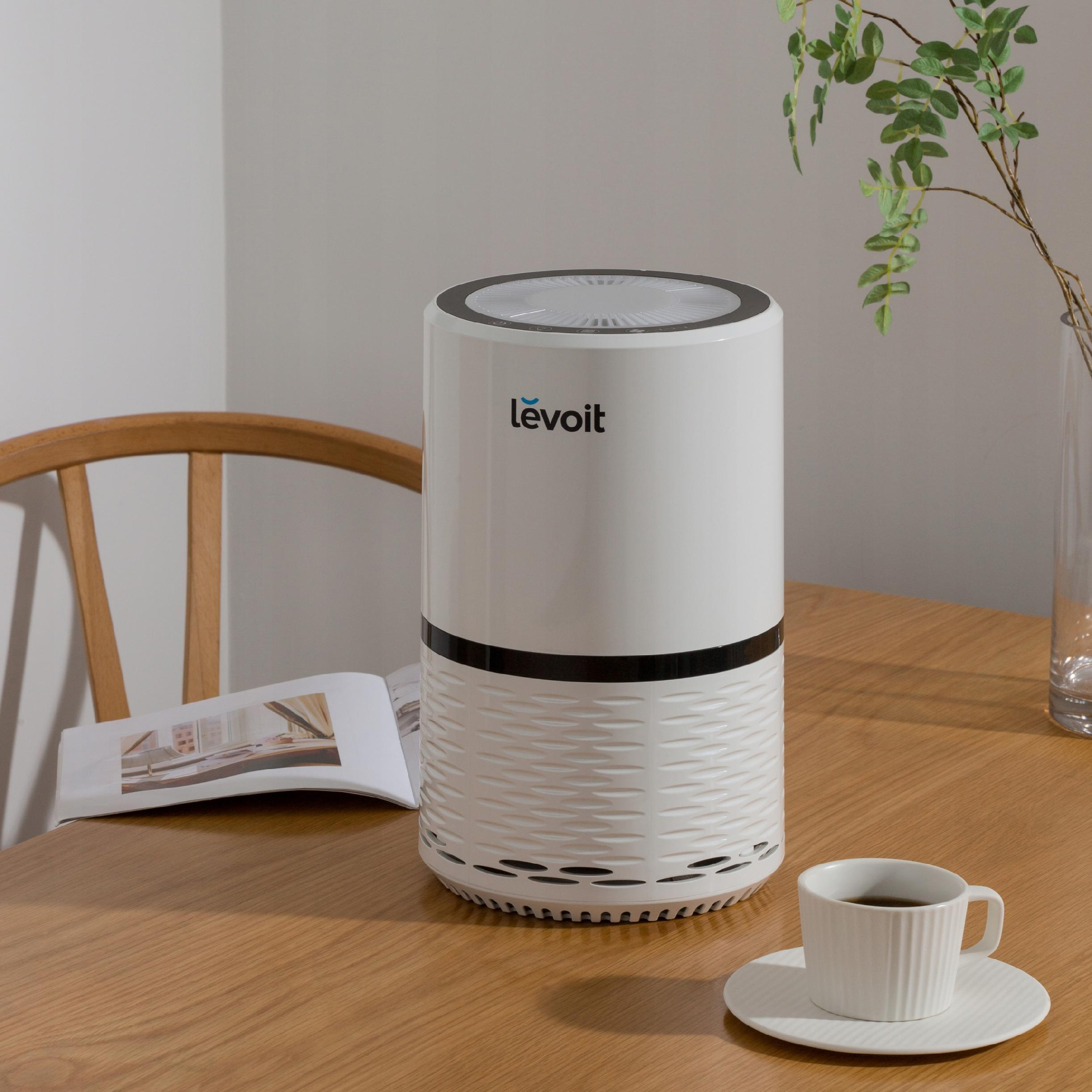 Biały oczyszczacz powietrza stoi na drewnianym stole. W pobliżu filiżanka kawy i czasopismo.