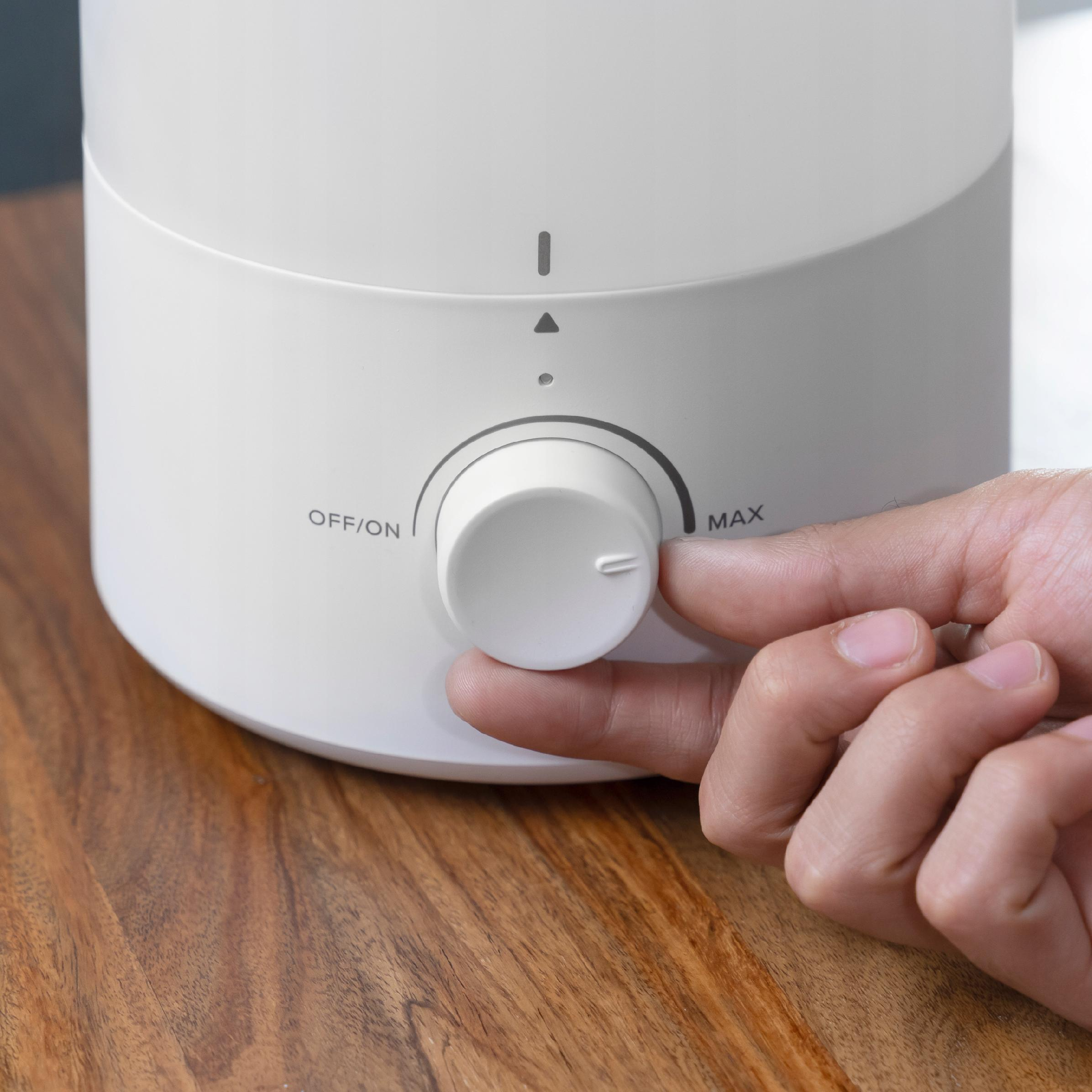 Ręka obraca pokrętło na białym nawilżaczu. Pokrętło ma oznaczenia 'OFF/ON' i 'MAX'. Tło jest drewniane.