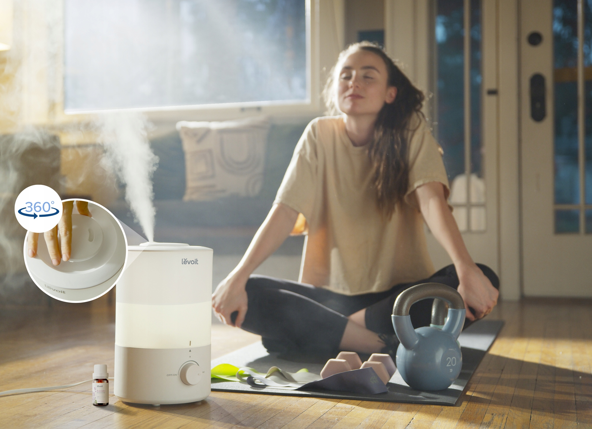 Kobieta medytuje w pobliżu nawilżacza. Nawilżacz jest biały. Na podłodze znajduje się kettlebell i inne przedmioty.