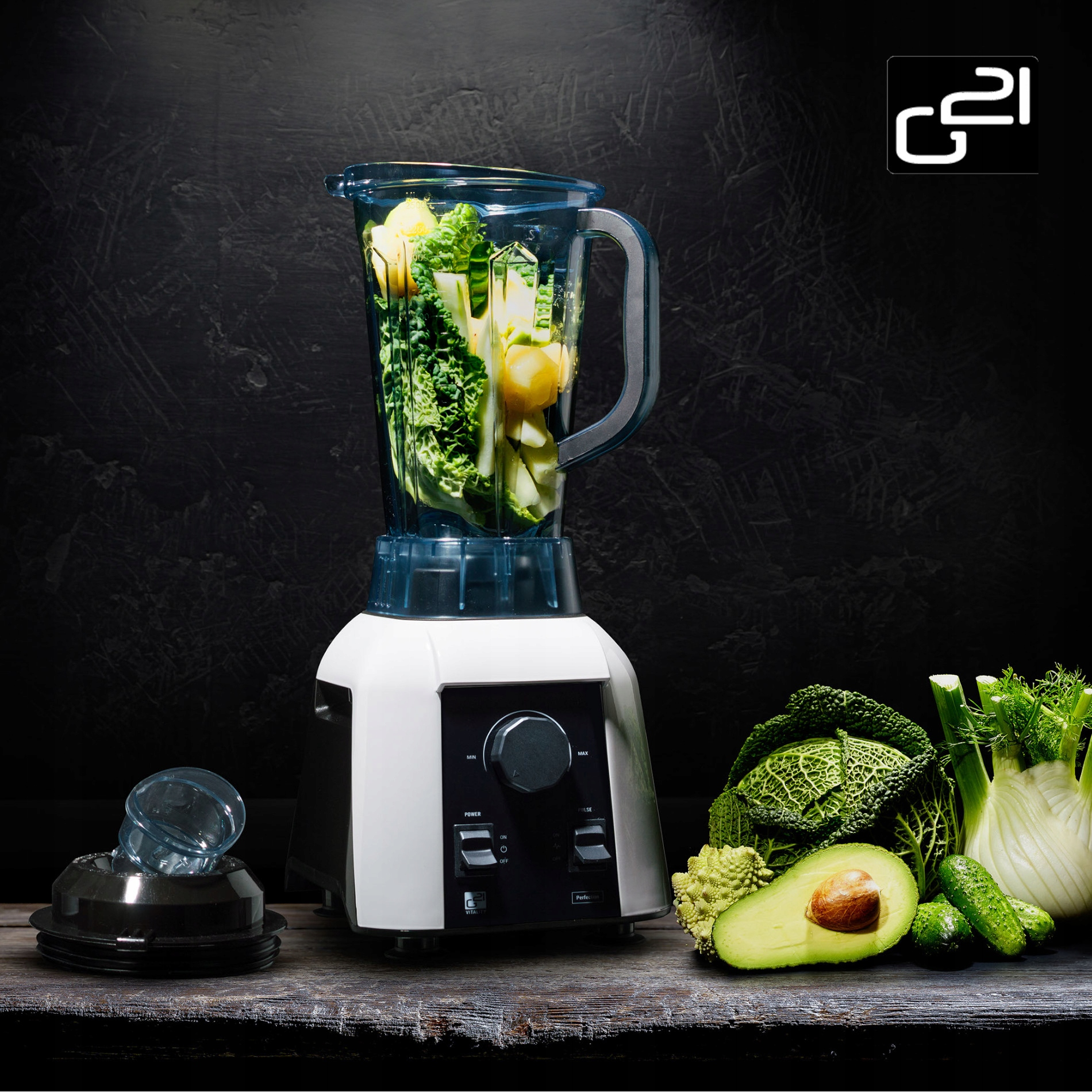 Blender z warzywami w środku. Niektóre warzywa są na stole. Tło jest czarne.