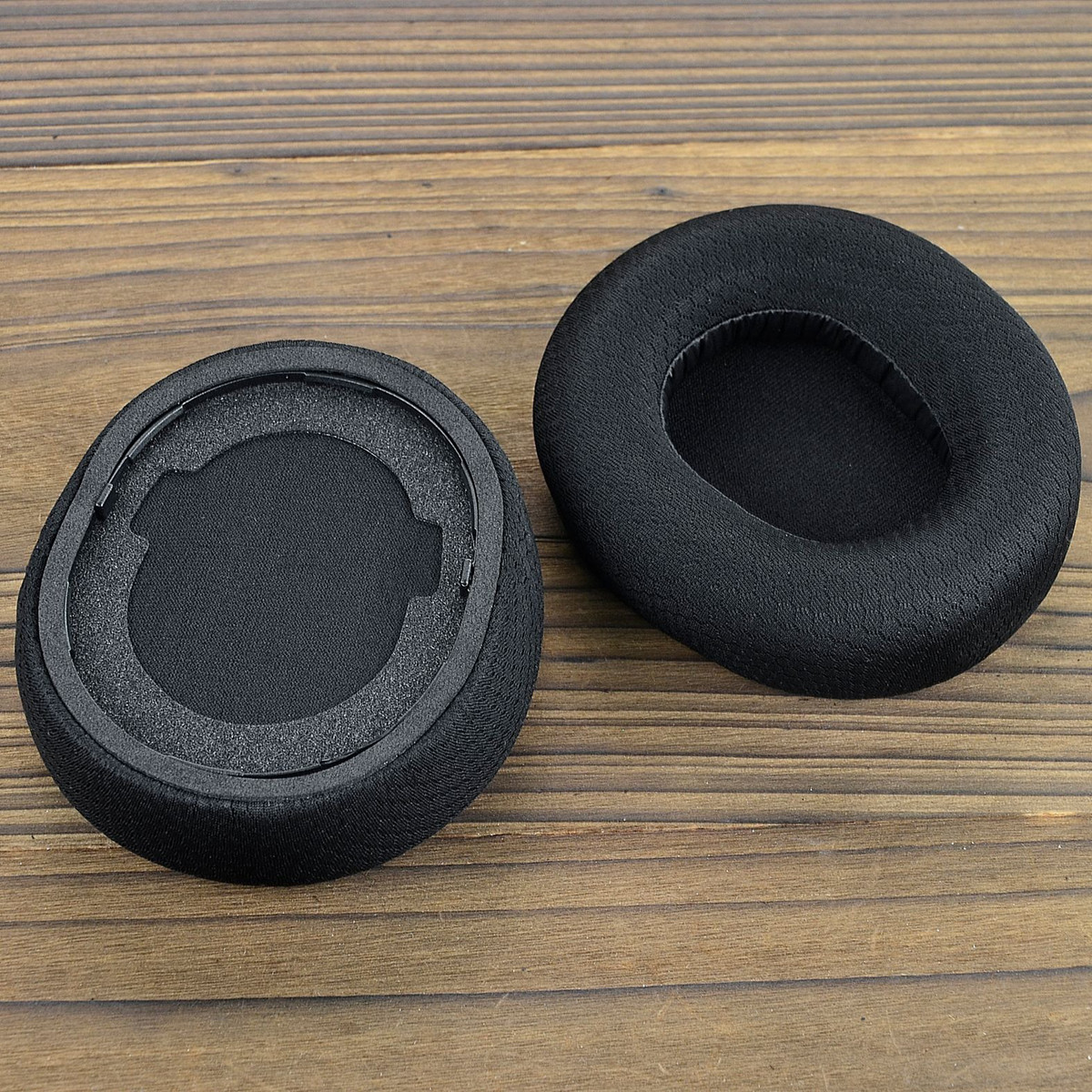 Due cuscinetti auricolari neri per cuffie su una superficie di legno. I cuscinetti hanno un materiale simile alla rete.