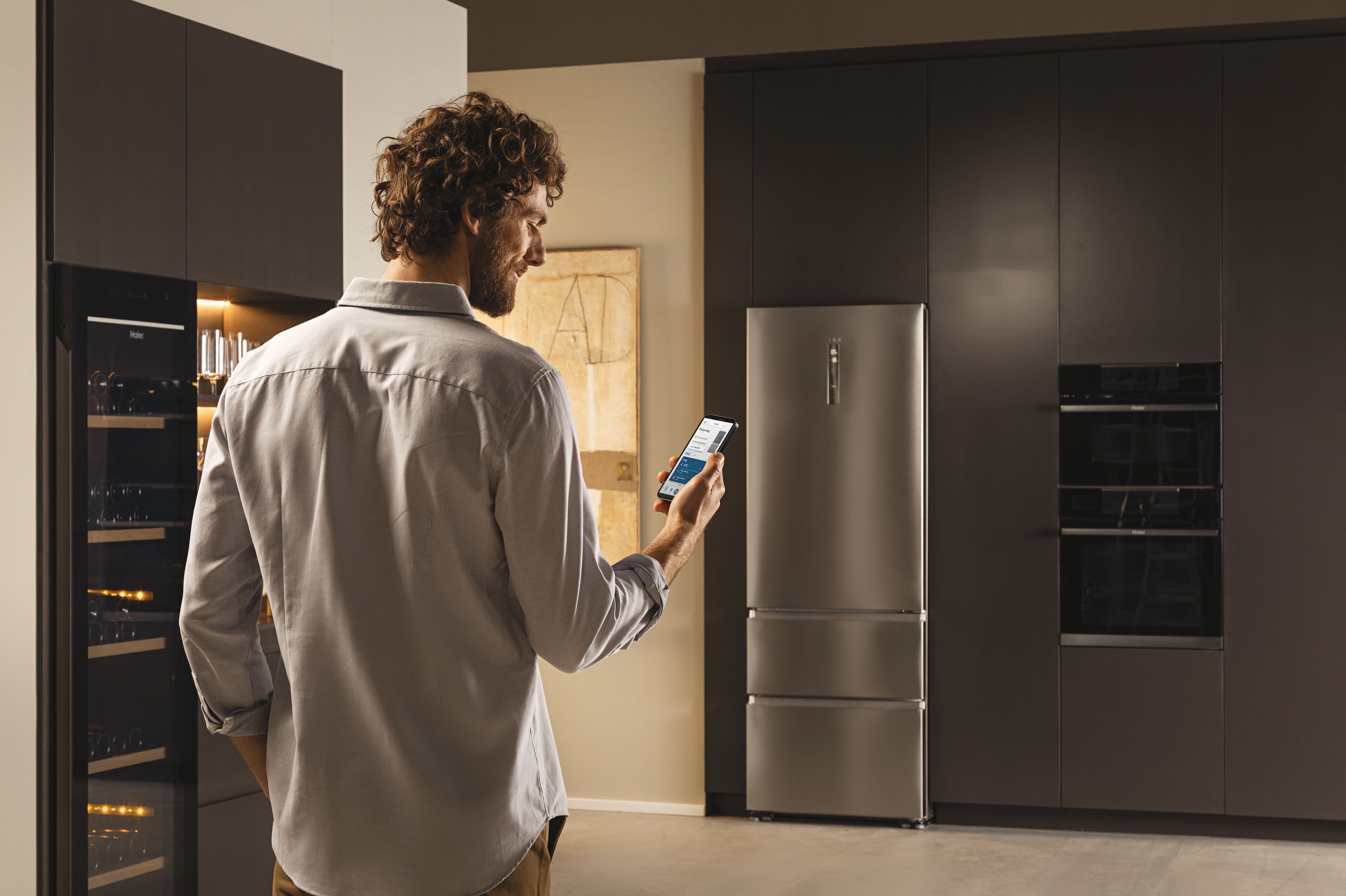 Un hombre usando un teléfono inteligente para controlar un refrigerador Haier en un entorno de cocina moderno.