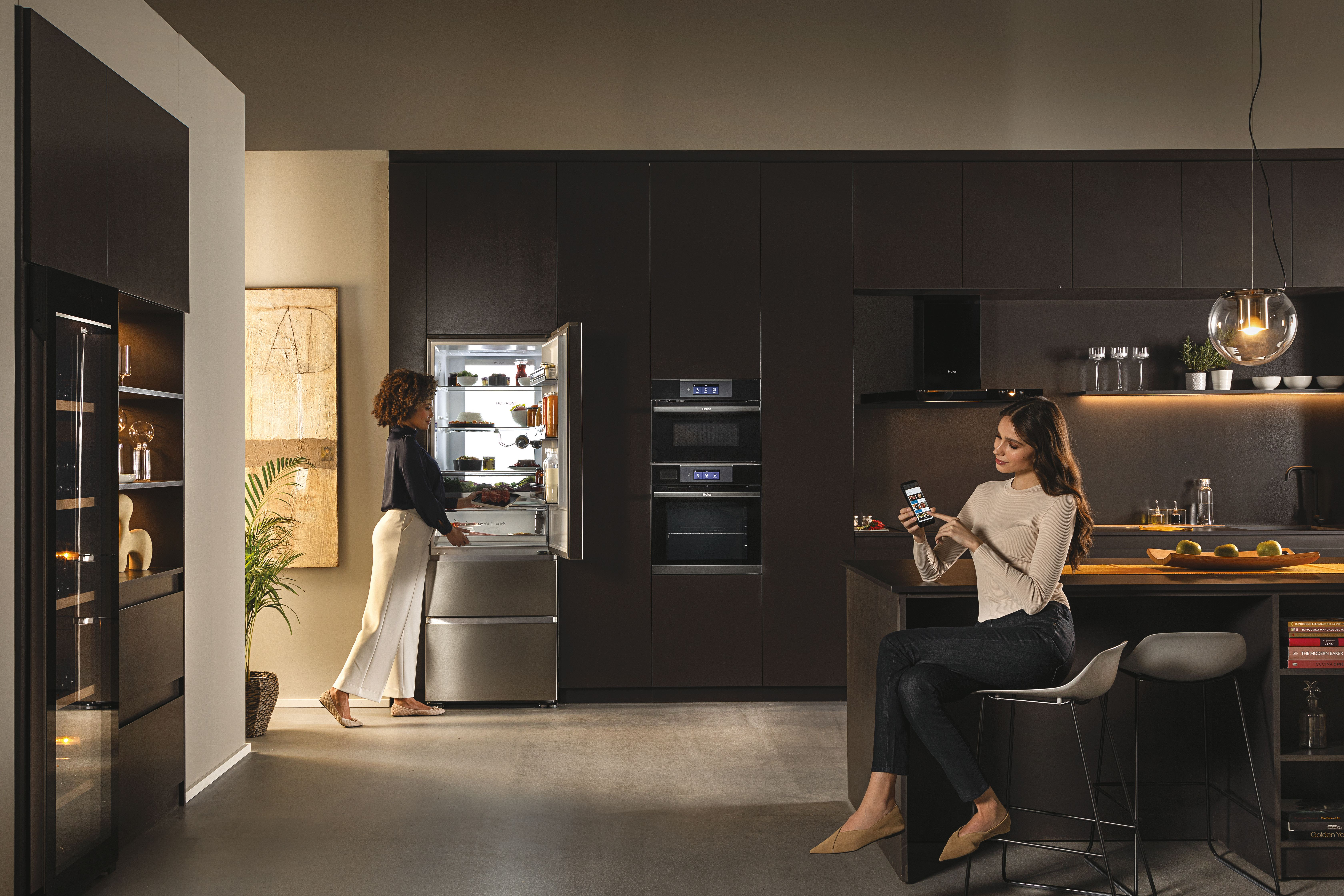 Escena de cocina moderna con dos mujeres, mostrando un refrigerador y tecnología inteligente para el hogar.