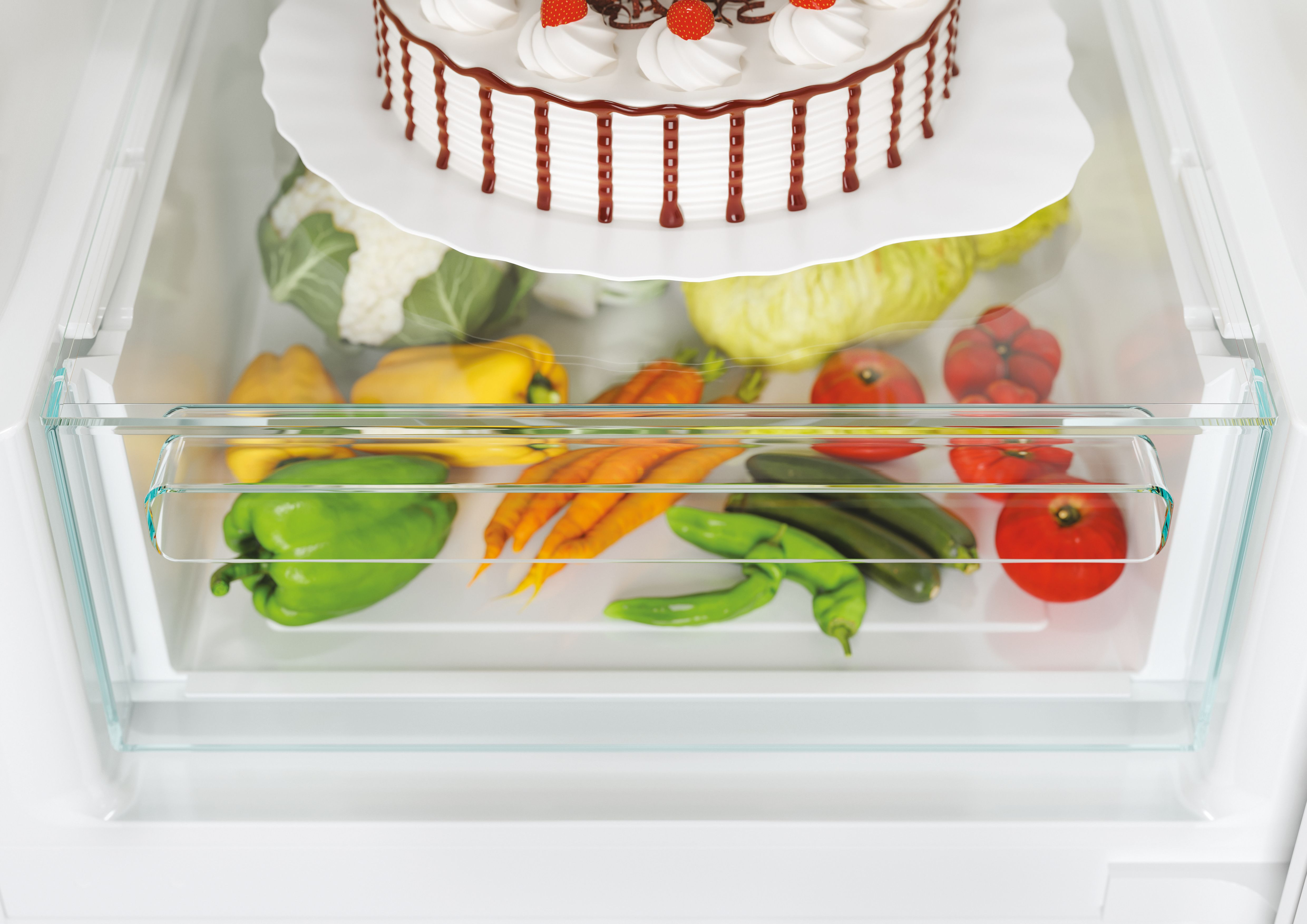 Primo piano di un cassetto del frigorifero pieno di verdure come carote, peperoni, pomodori e una torta sopra.