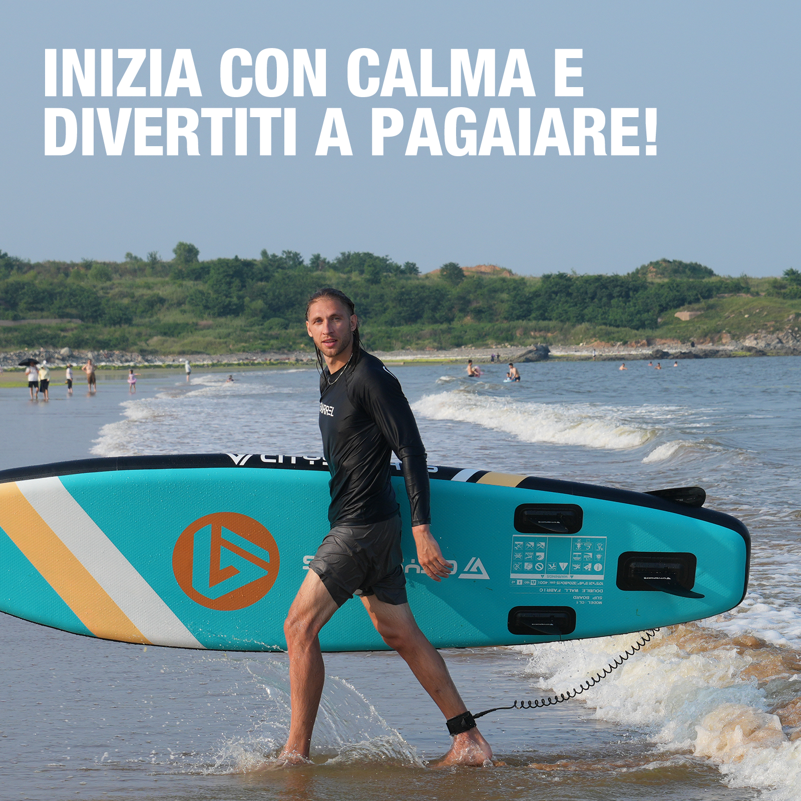 Un uomo porta una tavola da paddle sulla spiaggia. La tavola è blu e bianca. Ci sono persone sullo sfondo.
