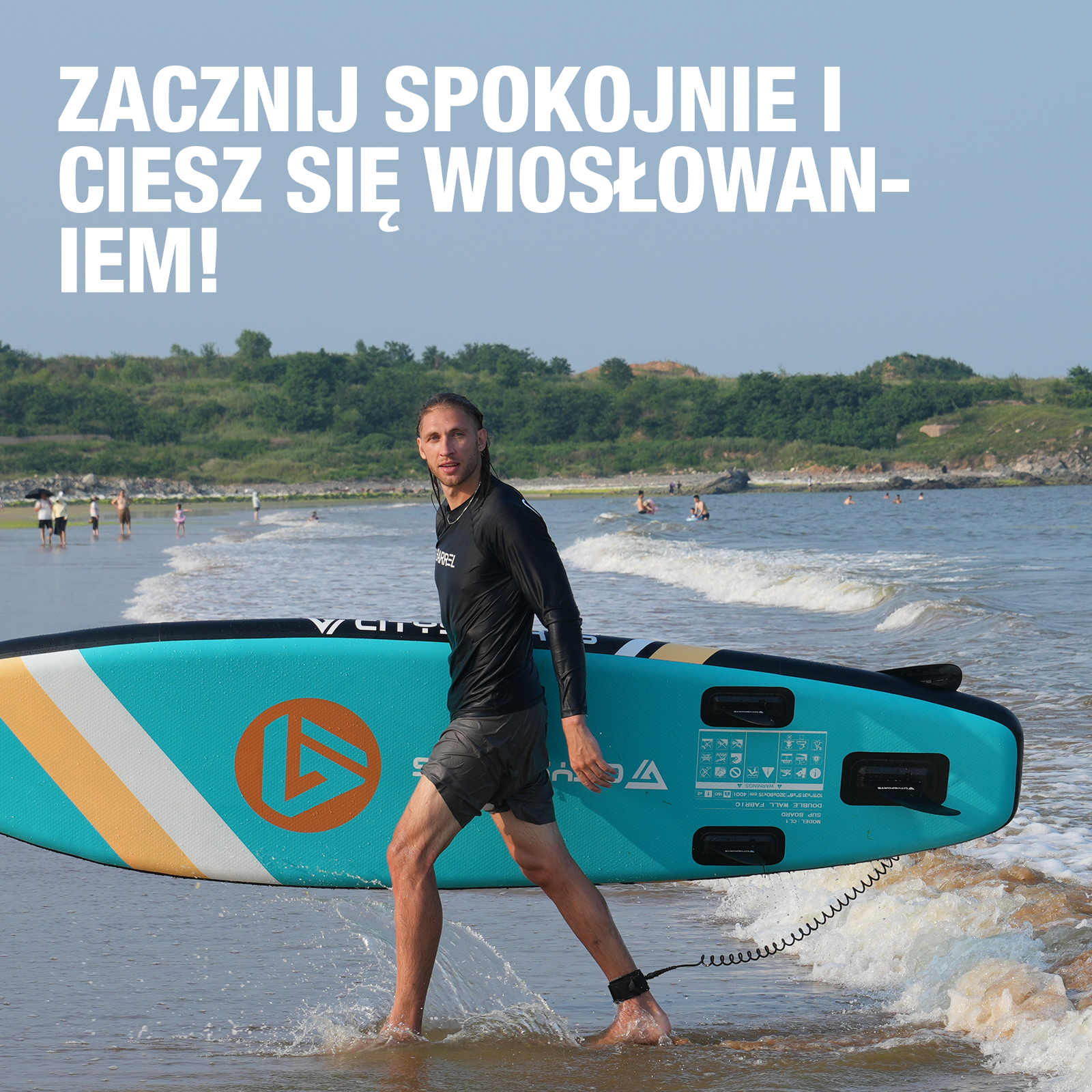 Mężczyzna niosący deskę SUP do morza. Deska jest niebiesko-żółta. Tekst mówi: Zacznij spokojnie i ciesz się wiosłowaniem!