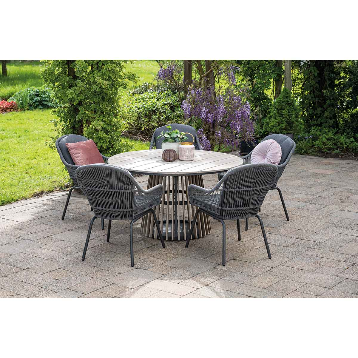A round outdoor table and four chairs are set on a stone patio with green foliage.