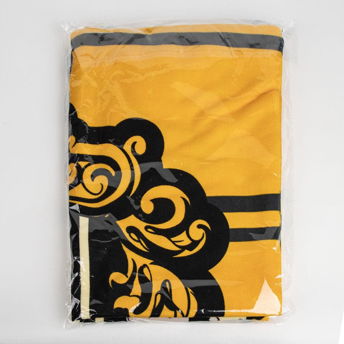 Toalla doblada en una bolsa de plástico. La toalla es dorada con rayas negras y un escudo en la esquina.