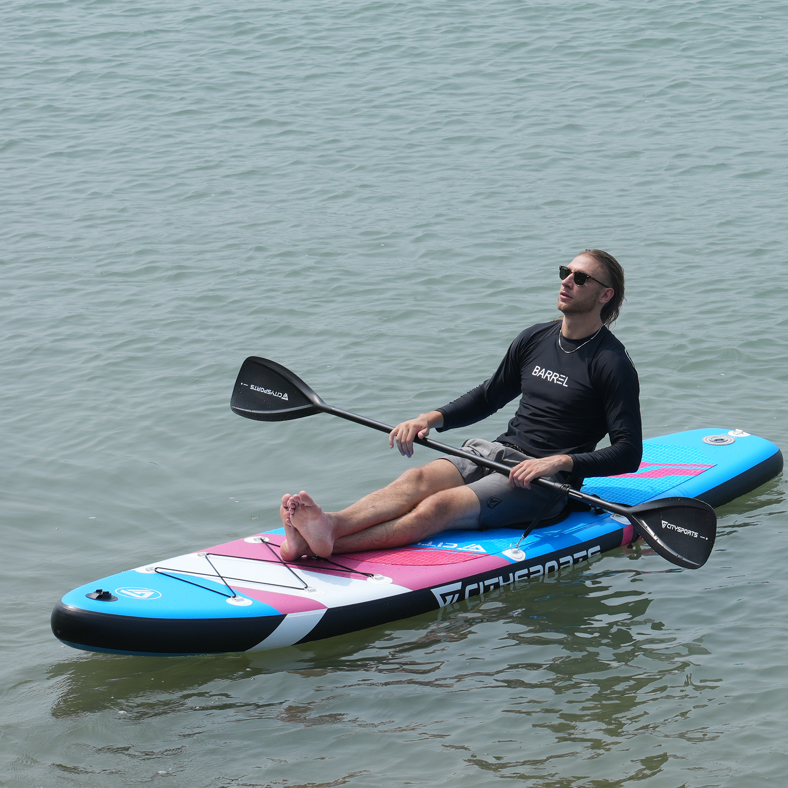 Mężczyzna w okularach przeciwsłonecznych siedzi na paddleboardzie, trzymając wiosło. Spokojna woda go otacza.