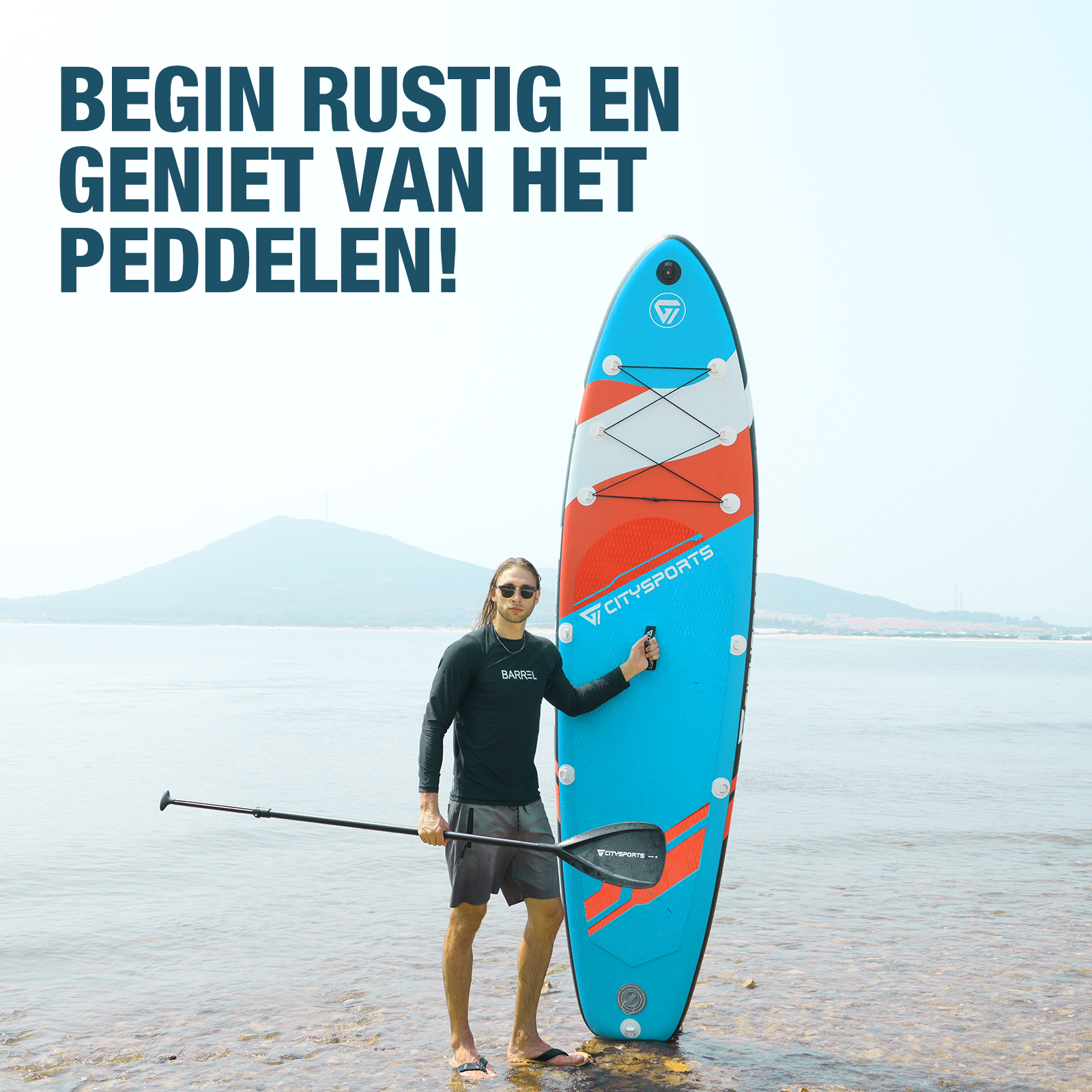 Homme debout avec un paddle, tenant une pagaie. Eau calme et montagnes en arrière-plan.