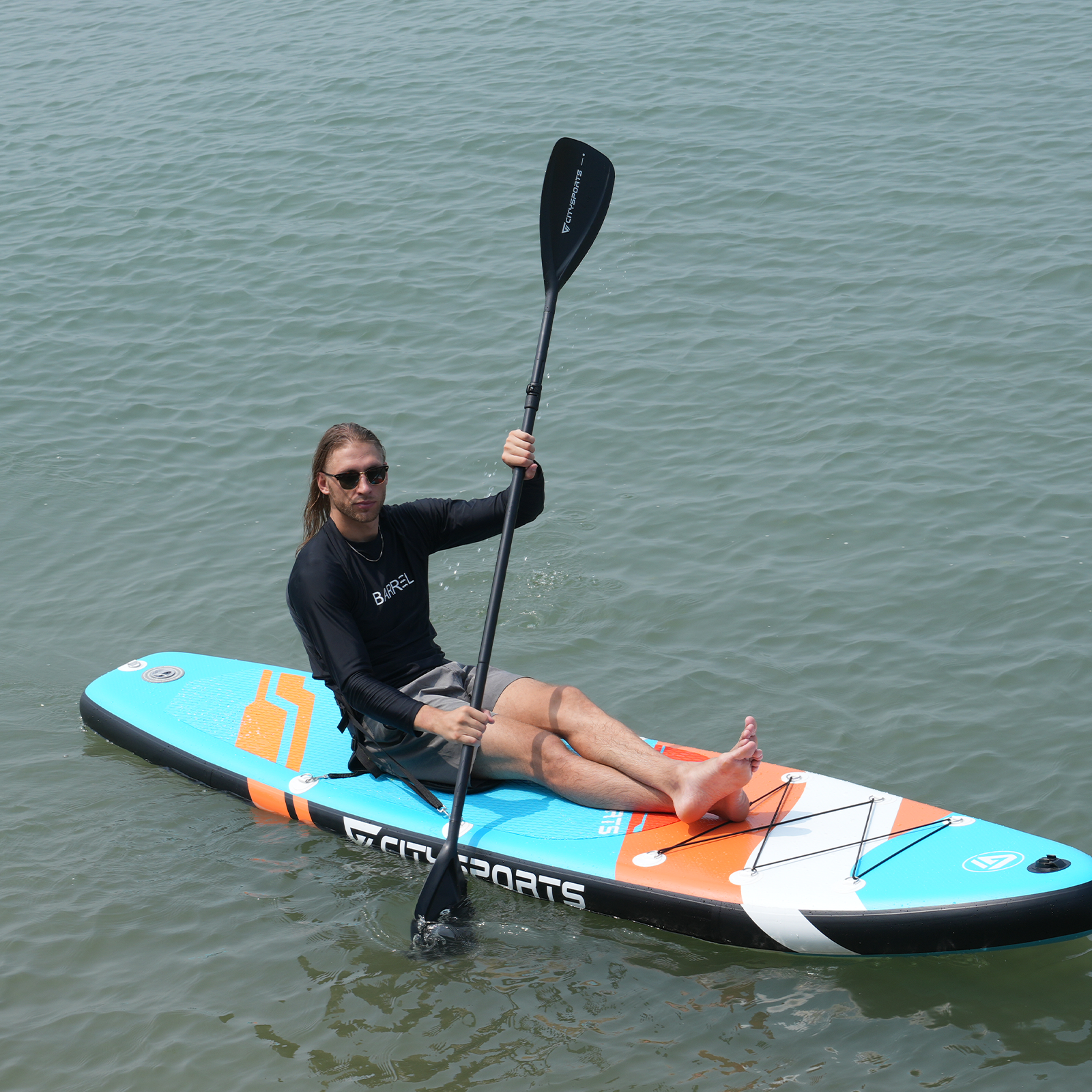 Mężczyzna w okularach przeciwsłonecznych siedzi na paddleboardzie, trzymając wiosło. Spokojna woda go otacza.