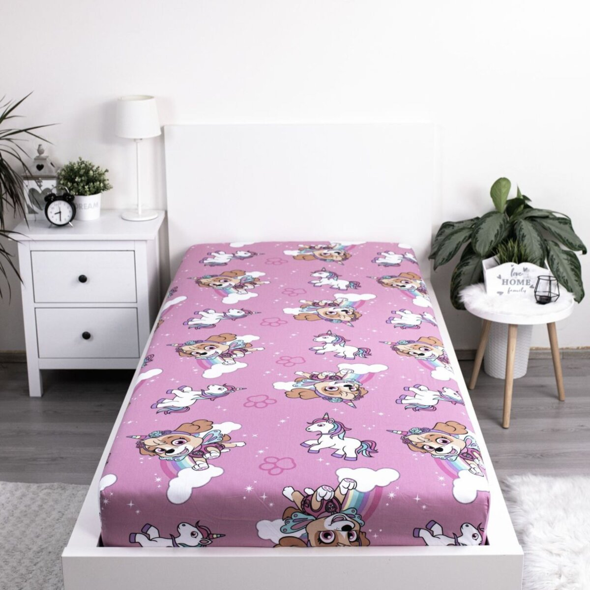 Ein pinkfarbenes Bettlaken mit Zeichentrickhunden und Einhörnern. Die Szene zeigt ein Bett, einen Nachttisch und eine Pflanze im Hintergrund.