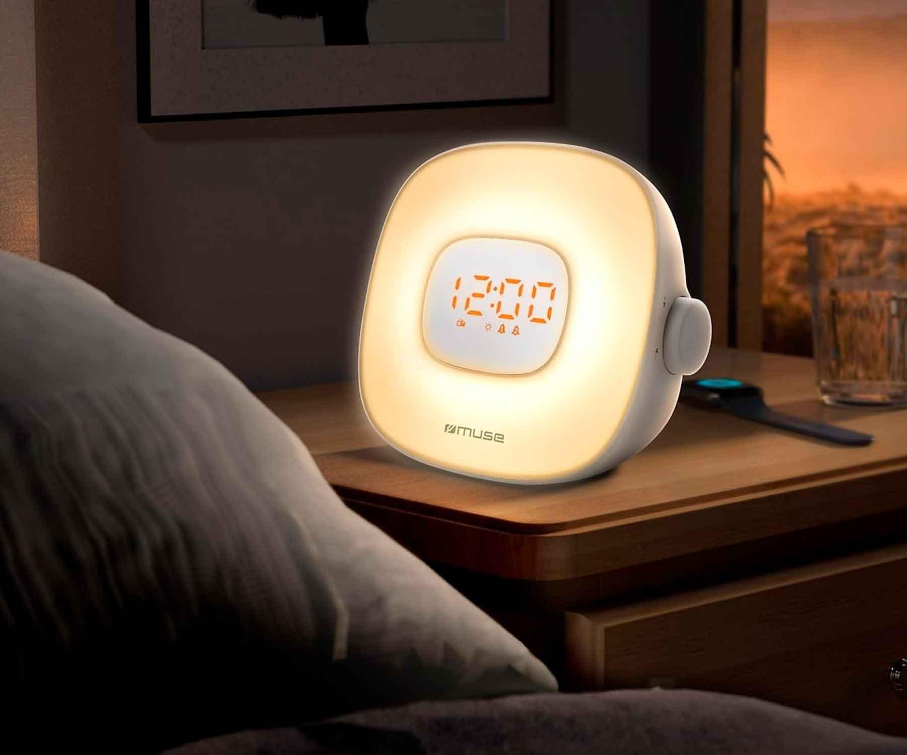Una lámpara de noche blanca que muestra la hora 12:00. Está sobre una mesita de noche de madera junto a una cama.