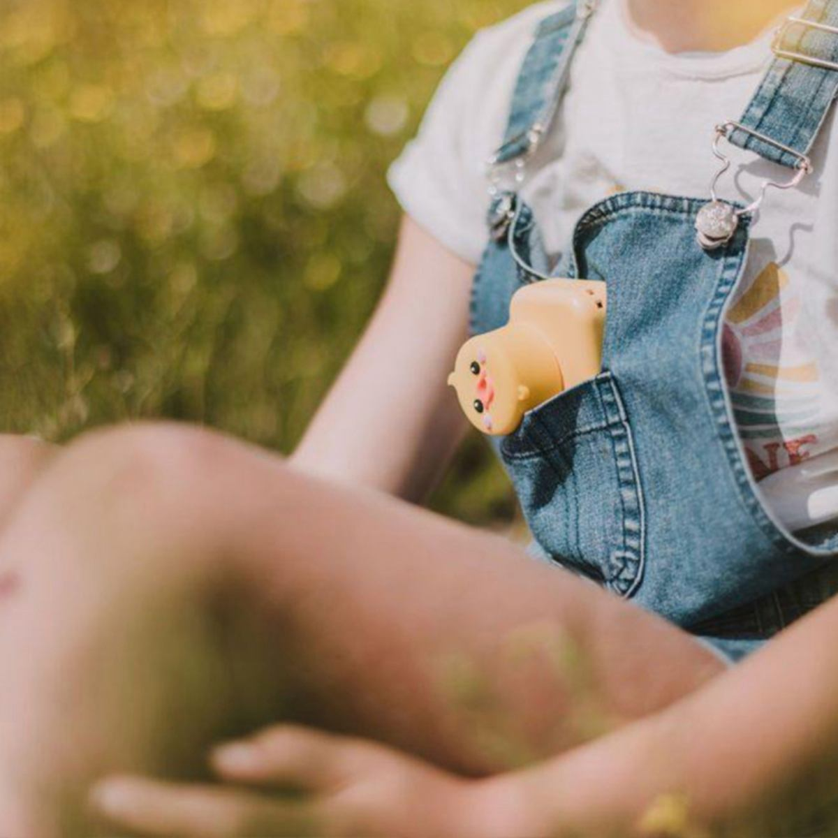 Ein Kind in Jeans-Latzhosen hält eine gelbe Enten-Kamera-Spielzeug in der Tasche. Draußen, sonnig, verschwommener Hintergrund.