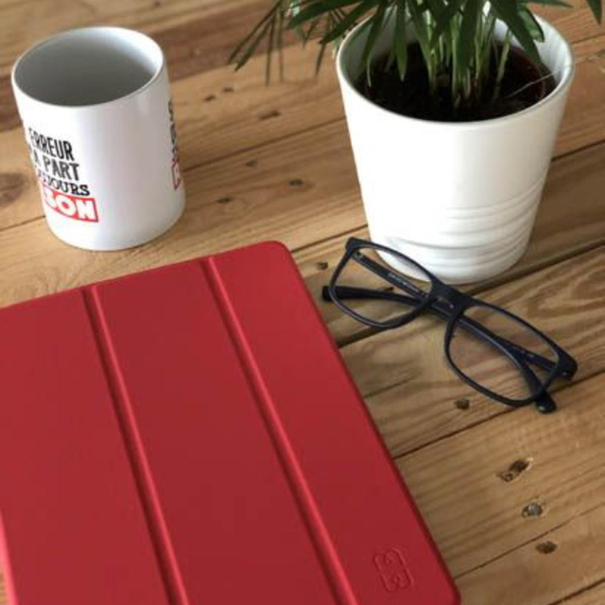 Una custodia rossa per tablet su un tavolo di legno, accanto a una tazza bianca e una pianta in un vaso bianco e occhiali.
